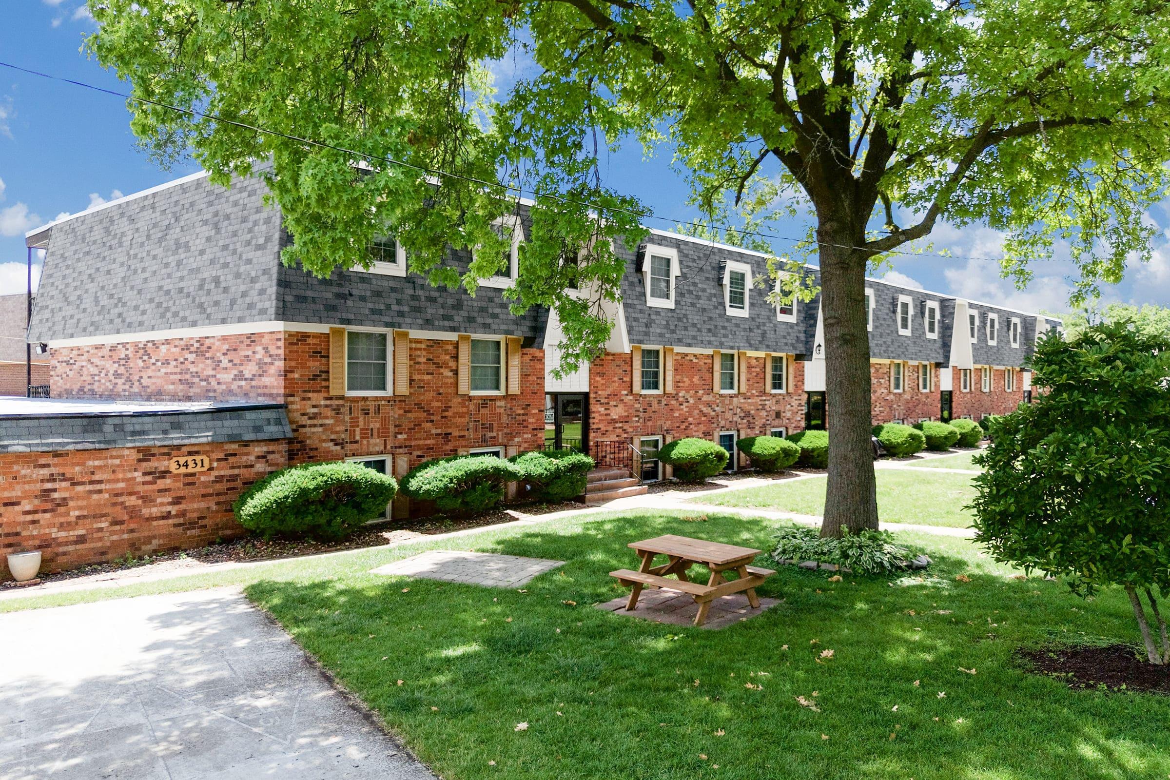 A view of a brick apartment building with gray accents, surrounded by green shrubs and neatly trimmed grass. A wooden picnic table is positioned on a pathway in front of the building, under the shade of a large tree. Blue sky and fluffy clouds are visible in the background.