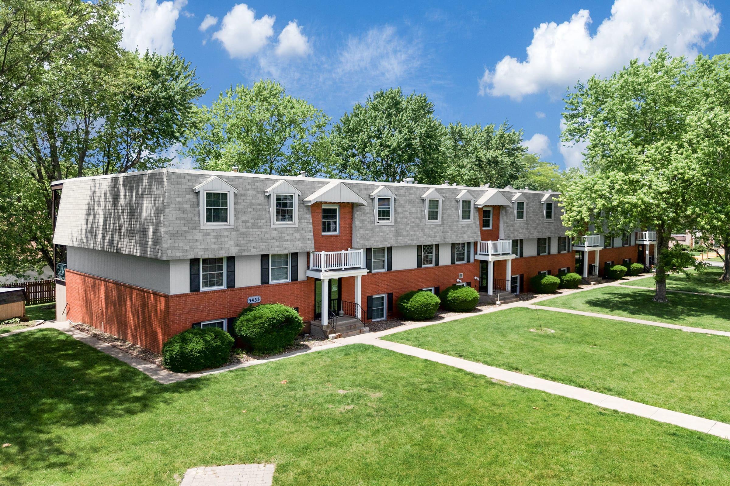 A multi-unit residential building with a red brick exterior and gray roof. The building features several balconies, surrounded by well-maintained green lawns and trees. The sky above is partly cloudy, adding a bright atmosphere to the scene.