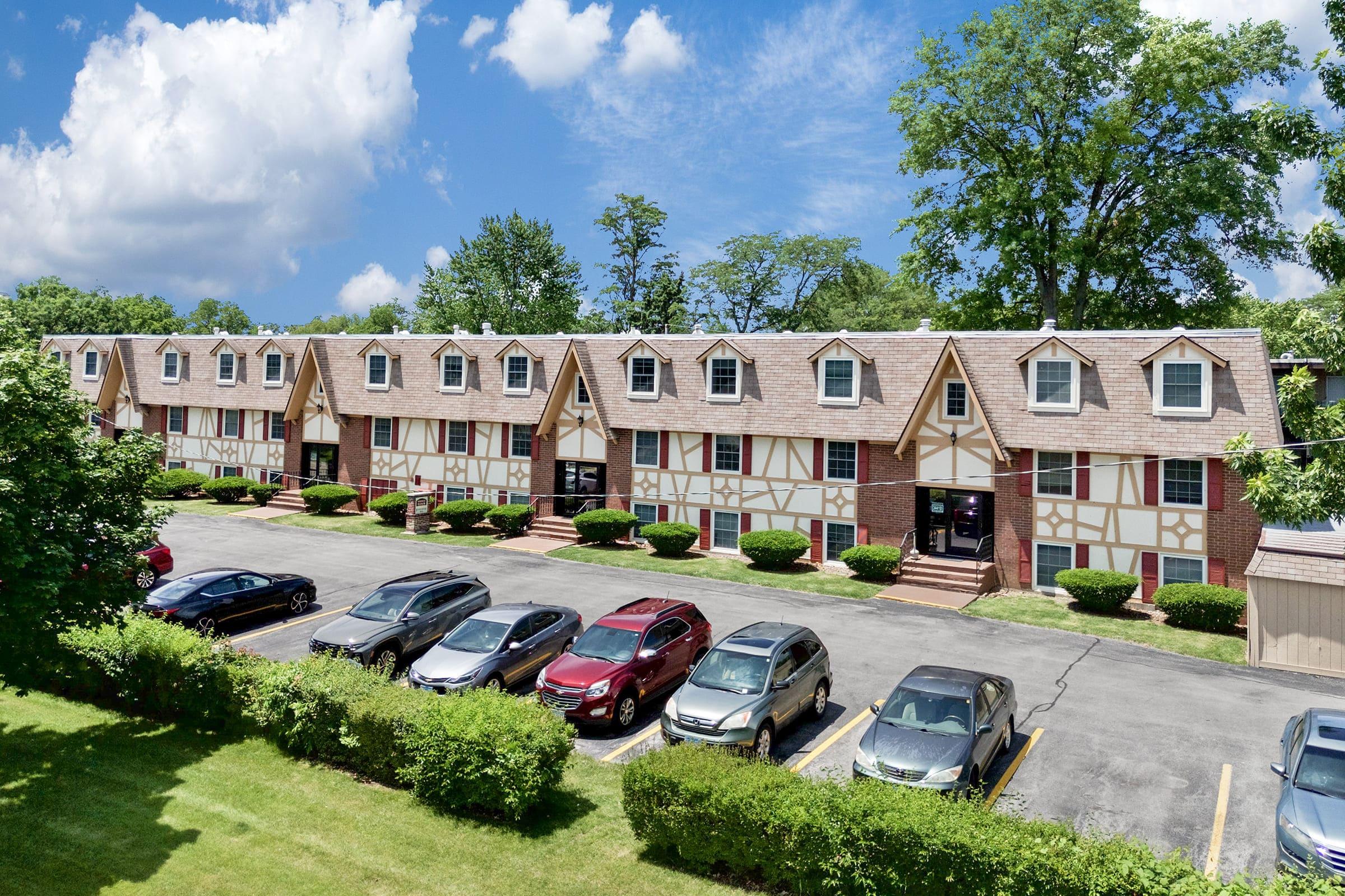 A view of a two-story, multi-unit residential building with a Tudor-style design, featuring a landscaped front lawn and a parking lot filled with various cars. The scene is set under a partly cloudy blue sky, surrounded by trees and greenery.