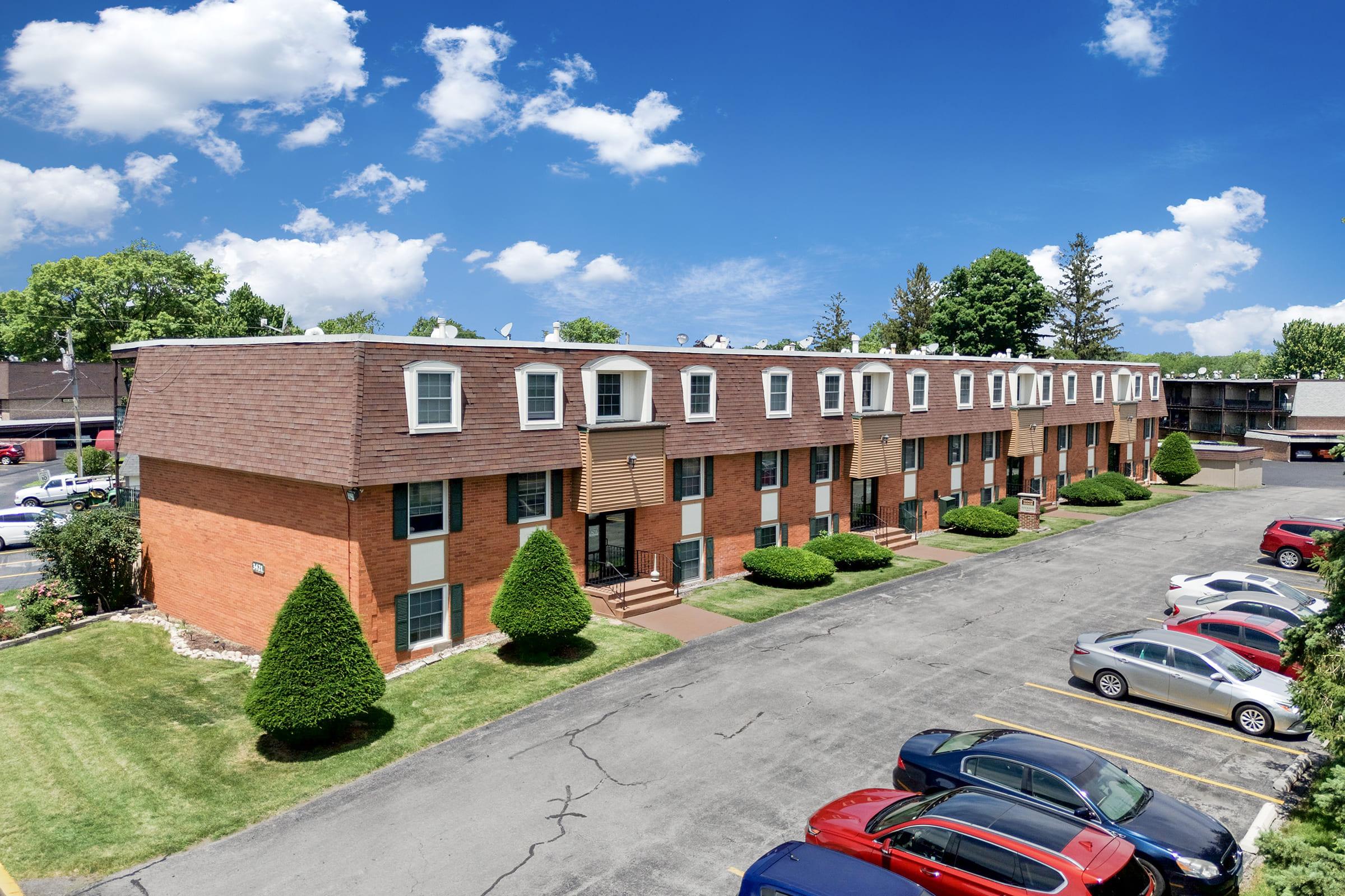 A row of brick apartment buildings with multiple windows and front doors, surrounded by well-manicured lawns and neatly trimmed bushes. The parking lot in the foreground has several cars parked. The sky is bright blue with fluffy white clouds, creating a cheerful atmosphere.
