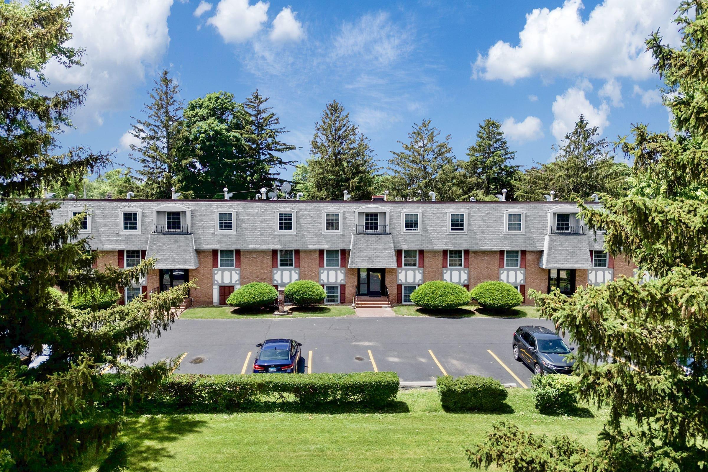 A view of a brick apartment building with multiple units, set against a blue sky with fluffy clouds. The building features grey roofs and white trim, surrounded by well-manicured shrubbery and trees. A parking lot in front has several parked cars.