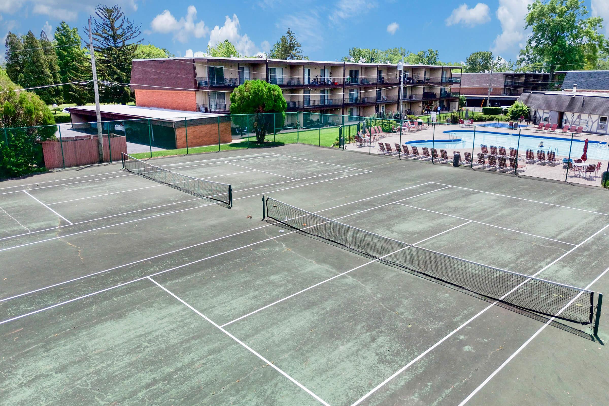 A view of several tennis courts in the foreground with a swimming pool and a multi-story building in the background. The courts are surrounded by green grass and trees. The sky is bright with fluffy clouds, indicating a sunny day. Sun loungers are arranged near the pool area.