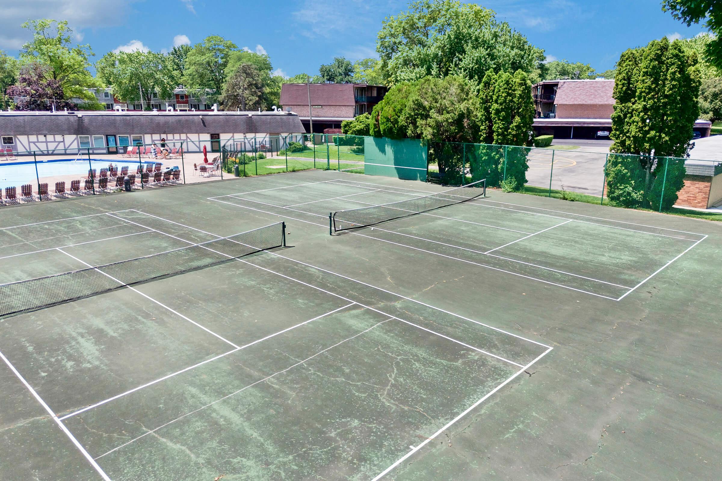 Aerial view of multiple tennis courts. The courts have faded green surfaces and white lines for marking. In the background, there is a swimming pool surrounded by lounge chairs and shaded trees. Nearby buildings are visible, providing a residential setting. The sky is clear with a few clouds.