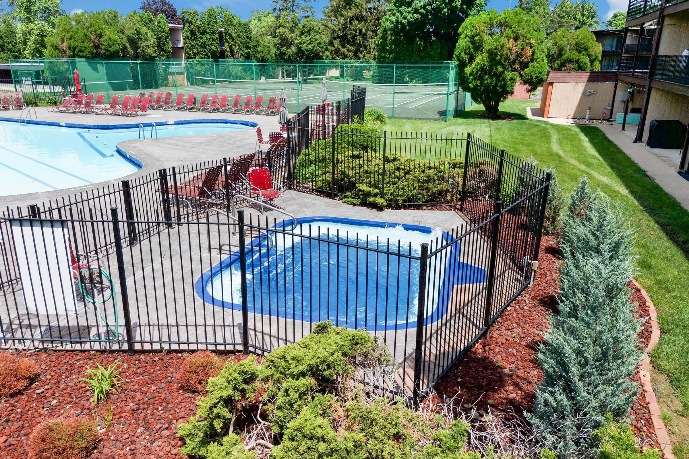 A view of a swimming pool area featuring a blue pool and a hot tub, surrounded by a black metal fence. Lush green grass and shrubs are visible, along with lounge chairs set up near the pool. In the background, tennis courts are partially visible, with a clear blue sky overhead.