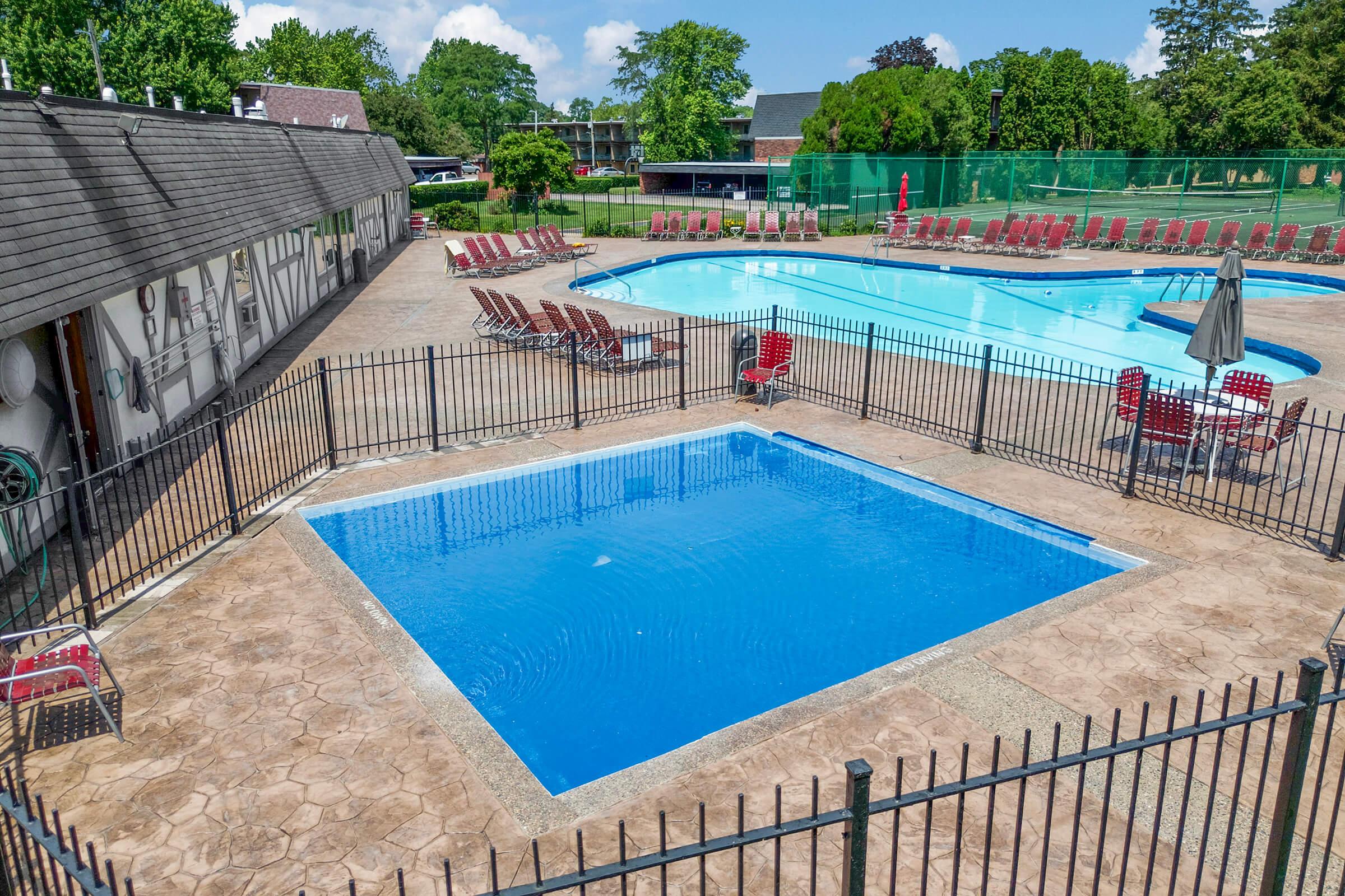 Aerial view of a swimming pool area featuring two pools: a larger blue pool surrounded by lounge chairs and a smaller empty pool. The area is enclosed by a black fence, with green trees and a clear sky in the background, creating a bright and inviting atmosphere.