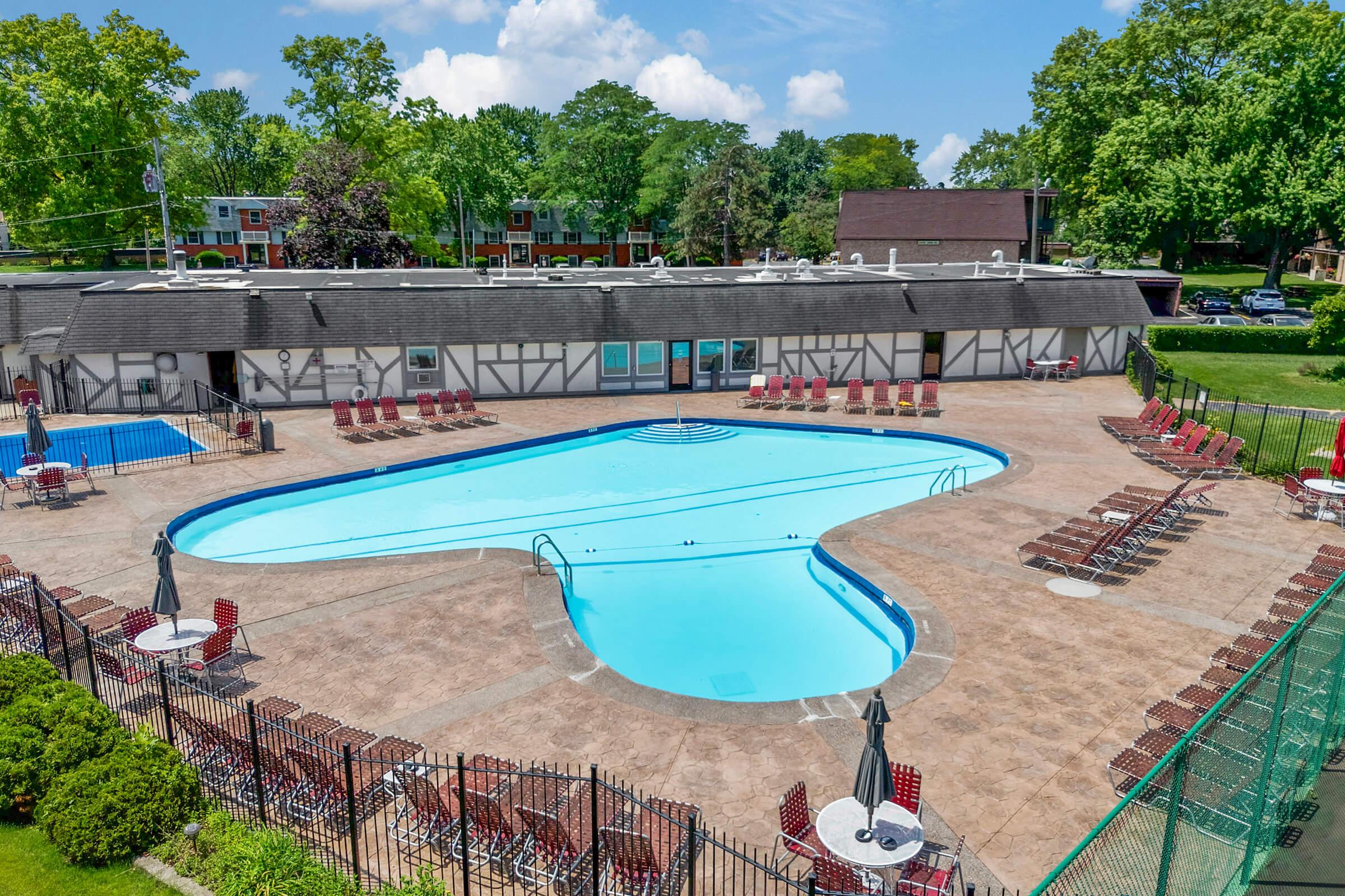 A large outdoor pool shaped like a kidney bean, surrounded by lounge chairs and umbrellas. The pool area is fenced and features a concrete deck. In the background, a building with a pitched roof and trees can be seen, under a blue sky with scattered clouds.