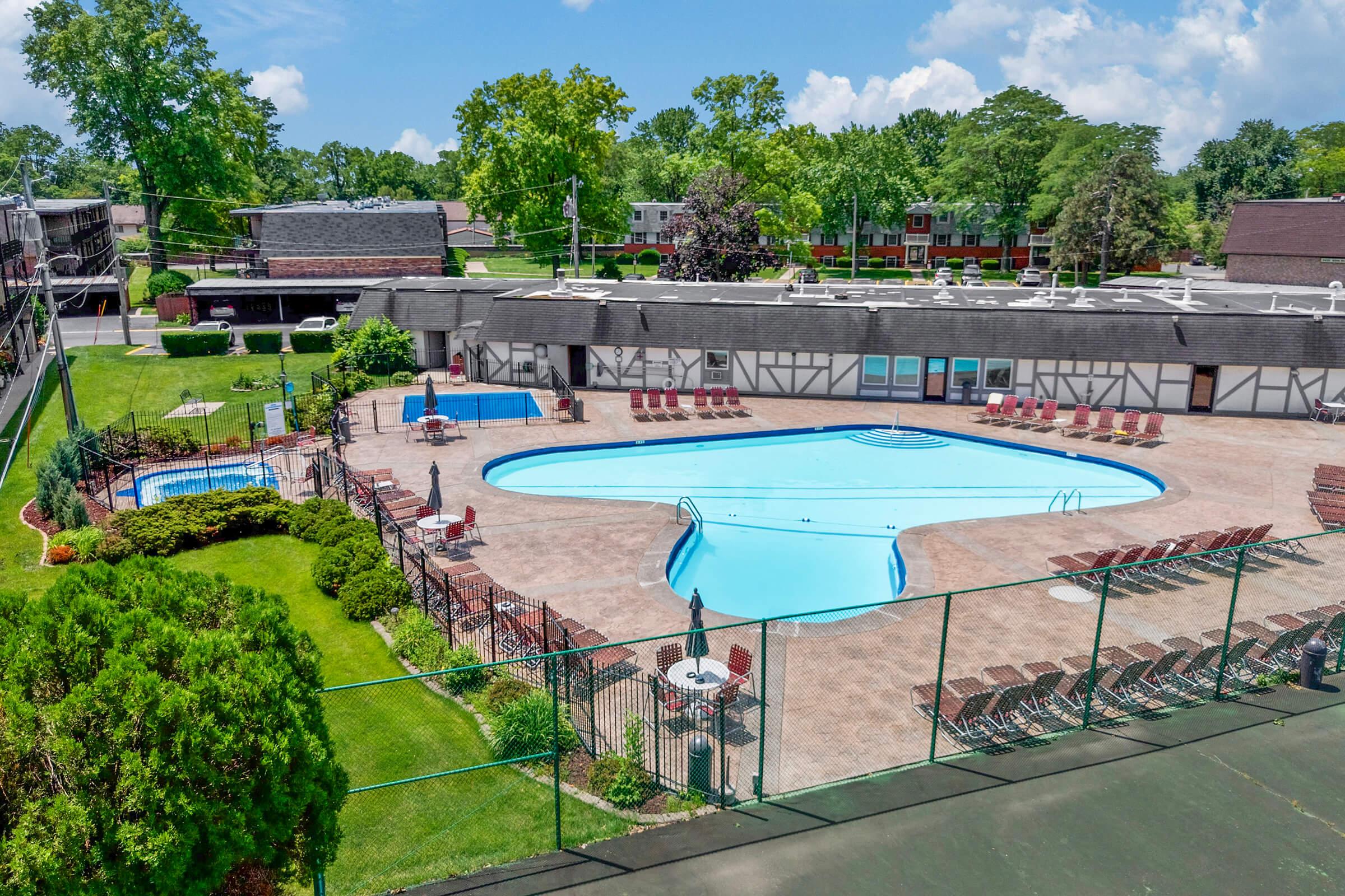 Aerial view of a hotel swimming pool area featuring a large, curved blue pool, a smaller pool, and numerous lounge chairs arranged around the deck. Lush green grass and trees surround the area, with buildings in the background under a bright blue sky with fluffy clouds.