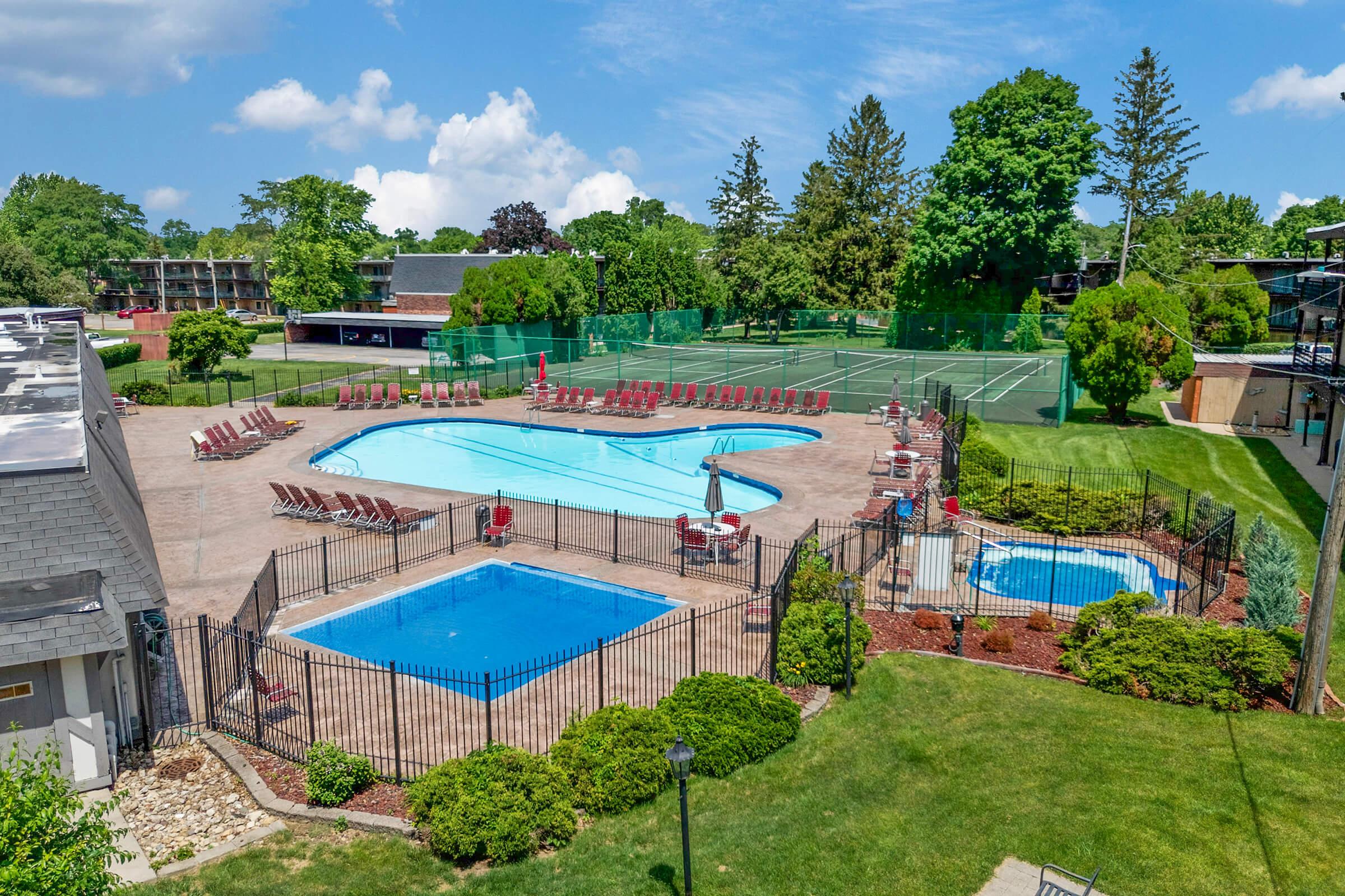 A spacious outdoor pool area featuring a large swimming pool, a smaller hot tub, and several lounge chairs arranged around the pool. Nearby, there are well-maintained tennis courts surrounded by greenery and trees under a clear blue sky.