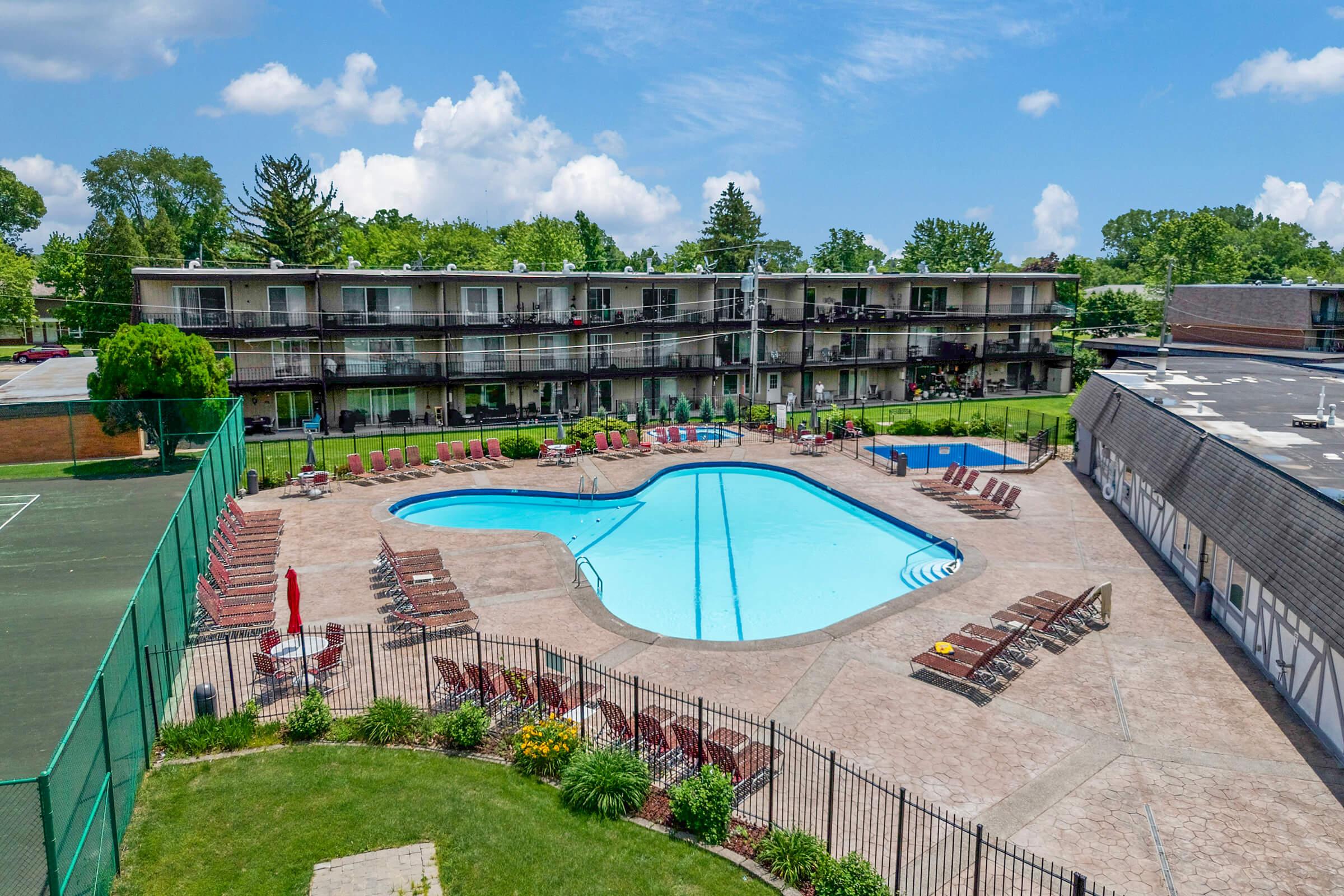 Aerial view of a hotel with a large, blue heart-shaped pool surrounded by lounge chairs. Green lawns and trees are in the background, with a tennis court on the left. The hotel building features multiple balconies and is situated under a bright, partly cloudy sky.