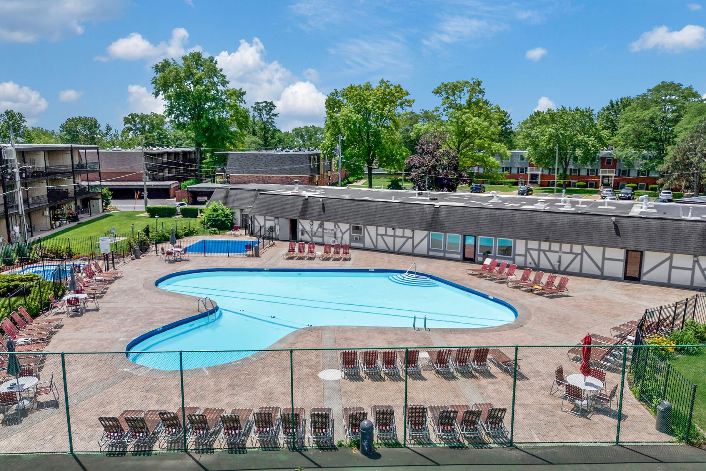 Aerial view of a heart-shaped swimming pool surrounded by lounge chairs, with a grassy area and trees in the background. The scene includes a tennis court and several adjacent buildings in a residential complex, all under a clear blue sky with fluffy white clouds.