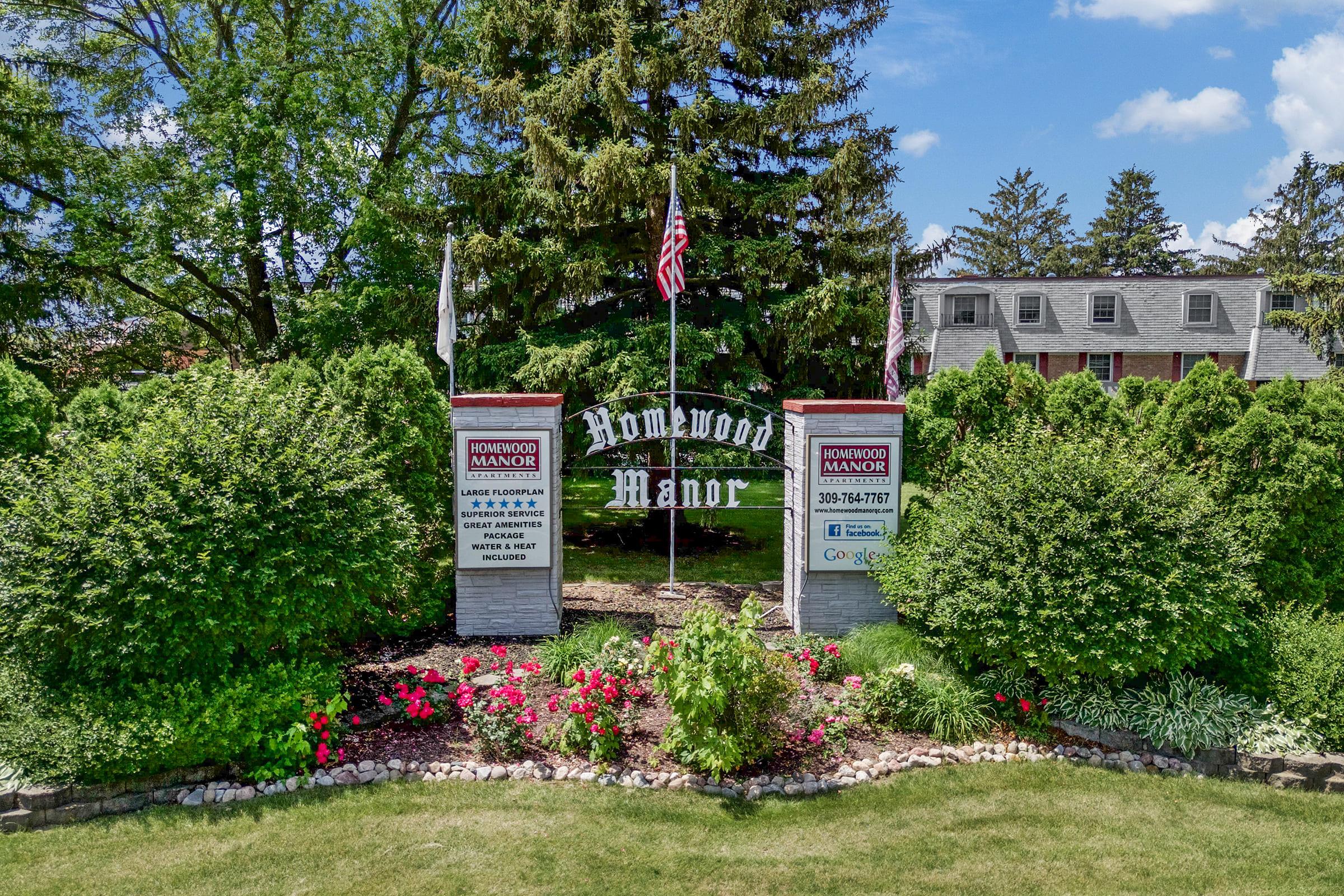 Signage for Homewood Manor surrounded by well-maintained greenery and colorful flower beds. Flags are displayed on both sides of the entrance, and a charming building is visible in the background under a bright blue sky.
