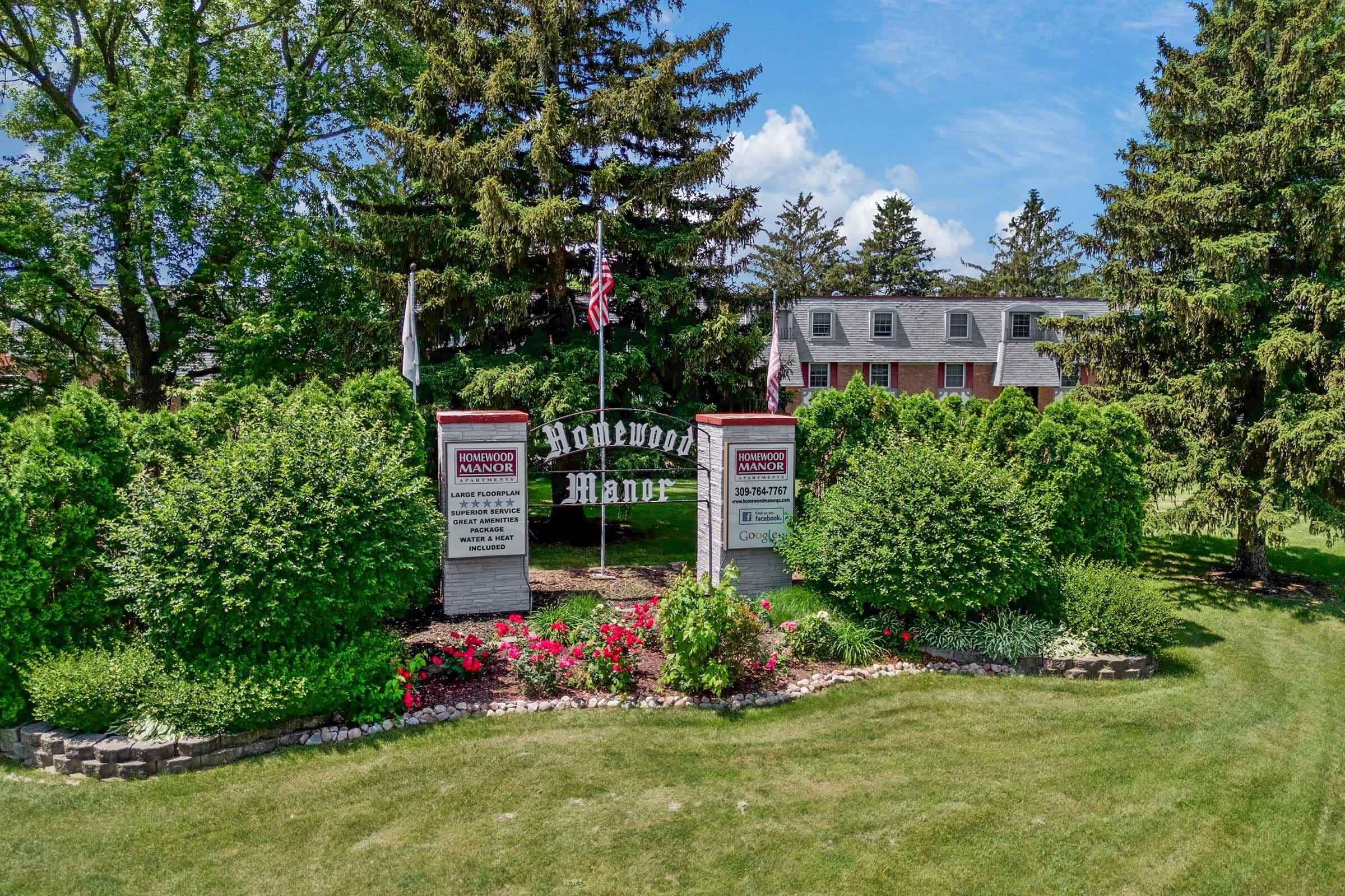 Signage for Homewood Manor surrounded by lush greenery and colorful flowers. Two large stone markers display the name "Homewood Manor" along with additional details. The background features a large building and several trees under a clear blue sky.