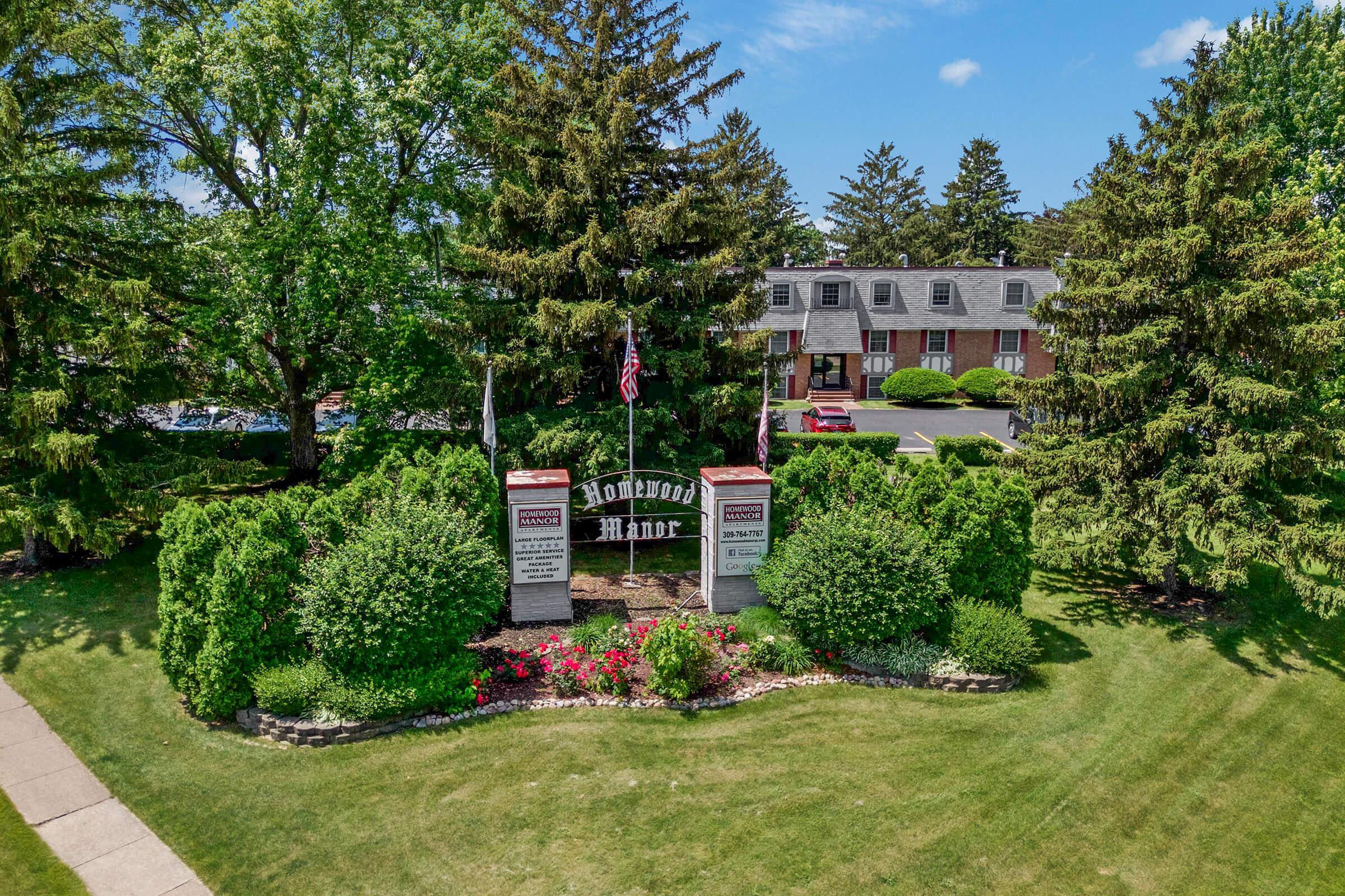 A landscaped entrance with a sign for "Homewood Manor," surrounded by neatly trimmed hedges, flowers, and tall trees. The background features a set of residential buildings and a clear blue sky. The scene is well-lit and conveys a welcoming atmosphere.