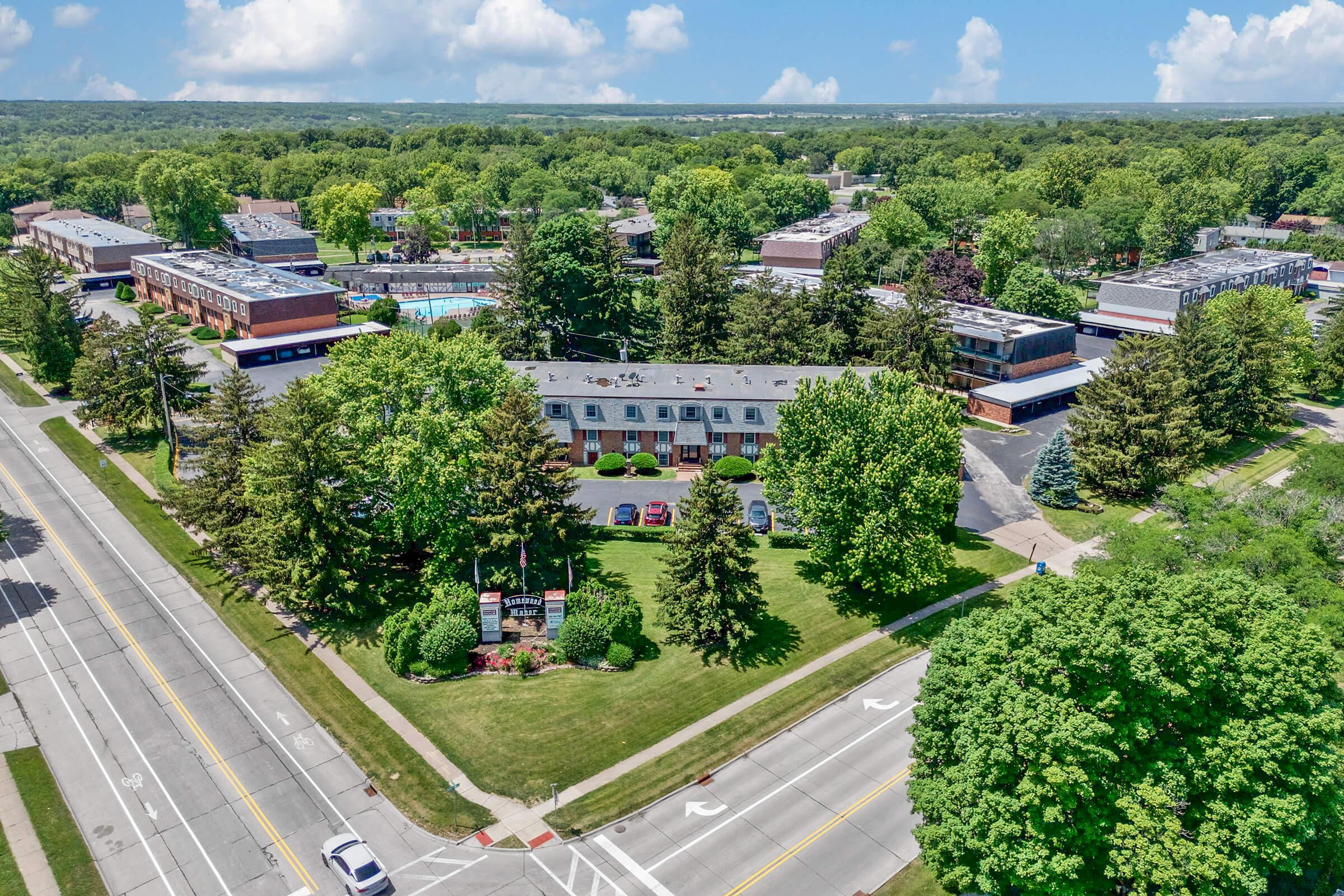 Aerial view of a hotel complex surrounded by lush greenery, featuring two-story buildings and a swimming pool area. The scene includes a well-maintained lawn and trees, with a road visible in the foreground. Bright blue sky with scattered clouds enhances the overall inviting atmosphere.