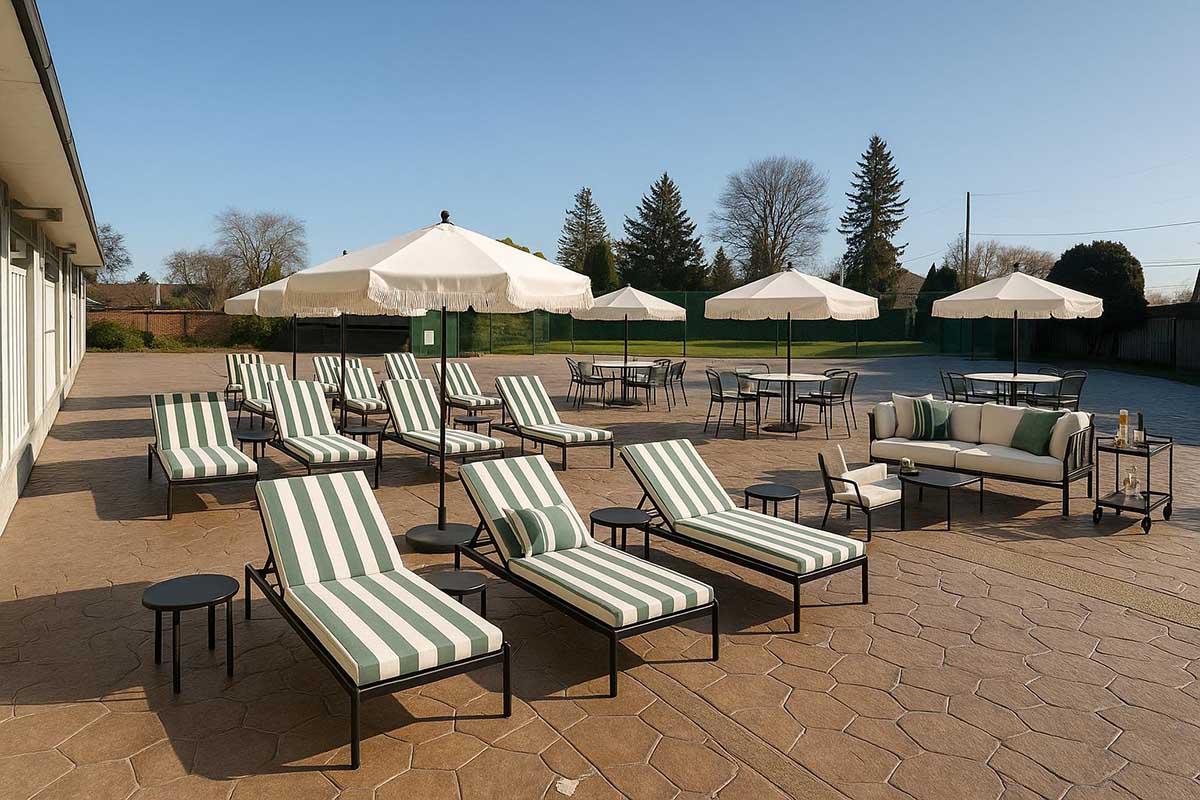 A spacious outdoor lounge area featuring multiple striped lounge chairs, small side tables, and a large seating arrangement. Several white umbrellas provide shade, and the floor is made of patterned stone. Trees and a clear sky are visible in the background, indicating a sunny day.