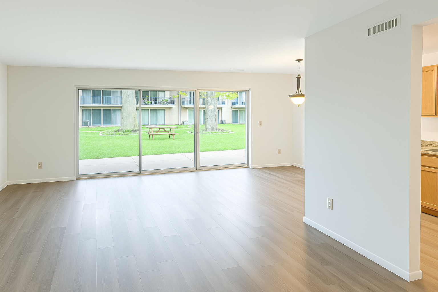 Bright and spacious living area featuring large sliding glass doors that open to a green outdoor space with trees and picnic tables. The room has light-colored flooring and neutral walls, creating an inviting atmosphere. Counter space and cabinetry are visible in the adjacent kitchen area.