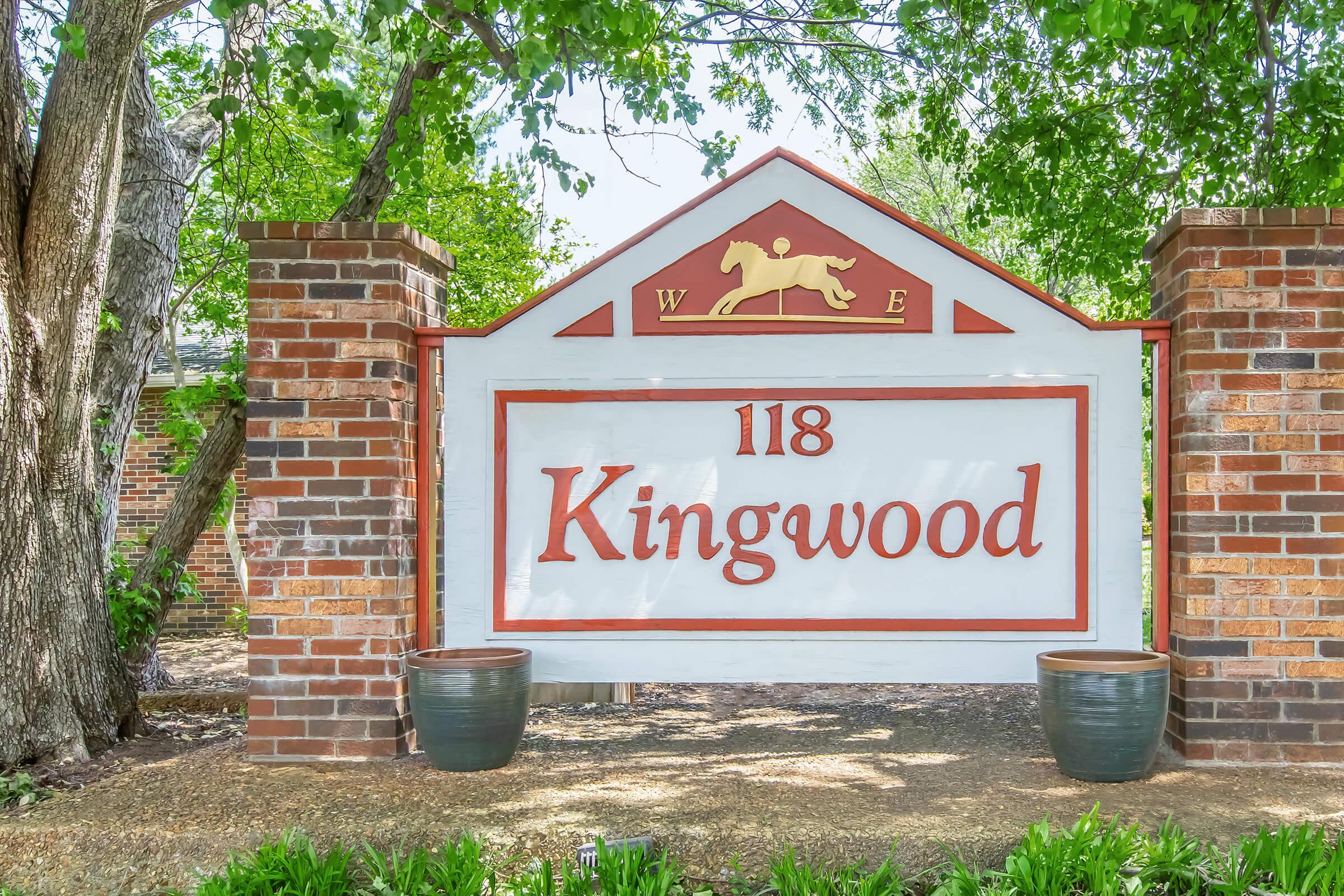 A brick sign featuring the text "118 Kingwood," with a horse silhouette above and decorative elements on either side. Surrounding the sign are green trees and potted plants, creating a welcoming entrance to a property.