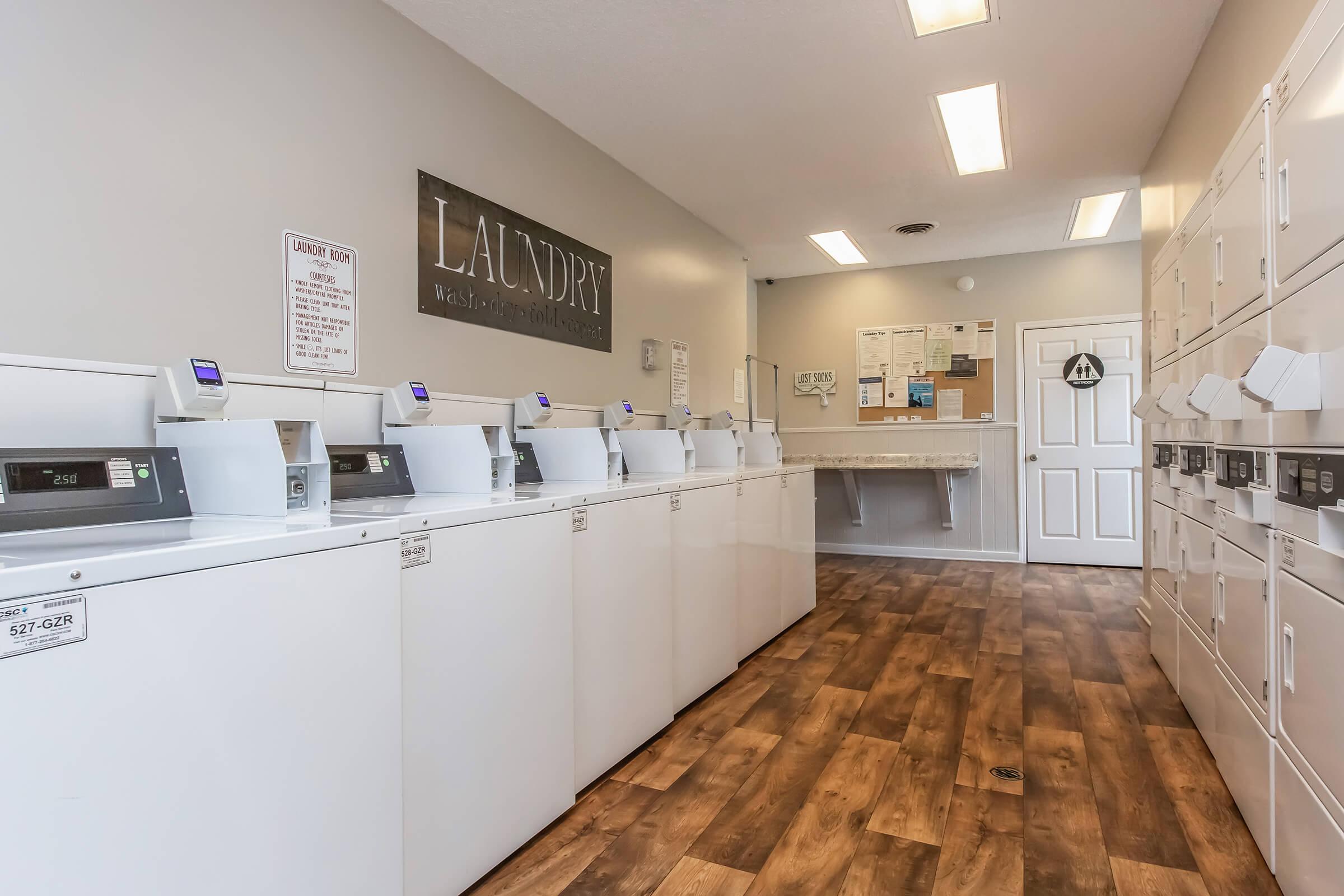 A clean, well-lit laundry room featuring several white washing machines and dryers arranged in a row. There is a folding area at the back, with bulletin boards and notices on the walls. The floor is made of wooden laminate, creating a warm, inviting atmosphere.