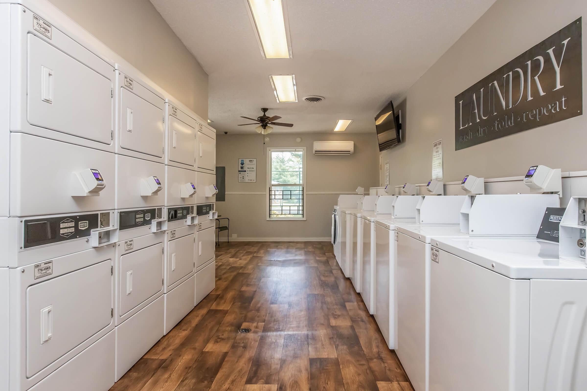 A clean and well-lit laundromat featuring rows of white washing machines and dryers along the walls. The floor is made of wooden planks, and there is a ceiling fan and a wall-mounted TV. A large sign reads "LAUNDRY" on one wall, with a window allowing natural light in.