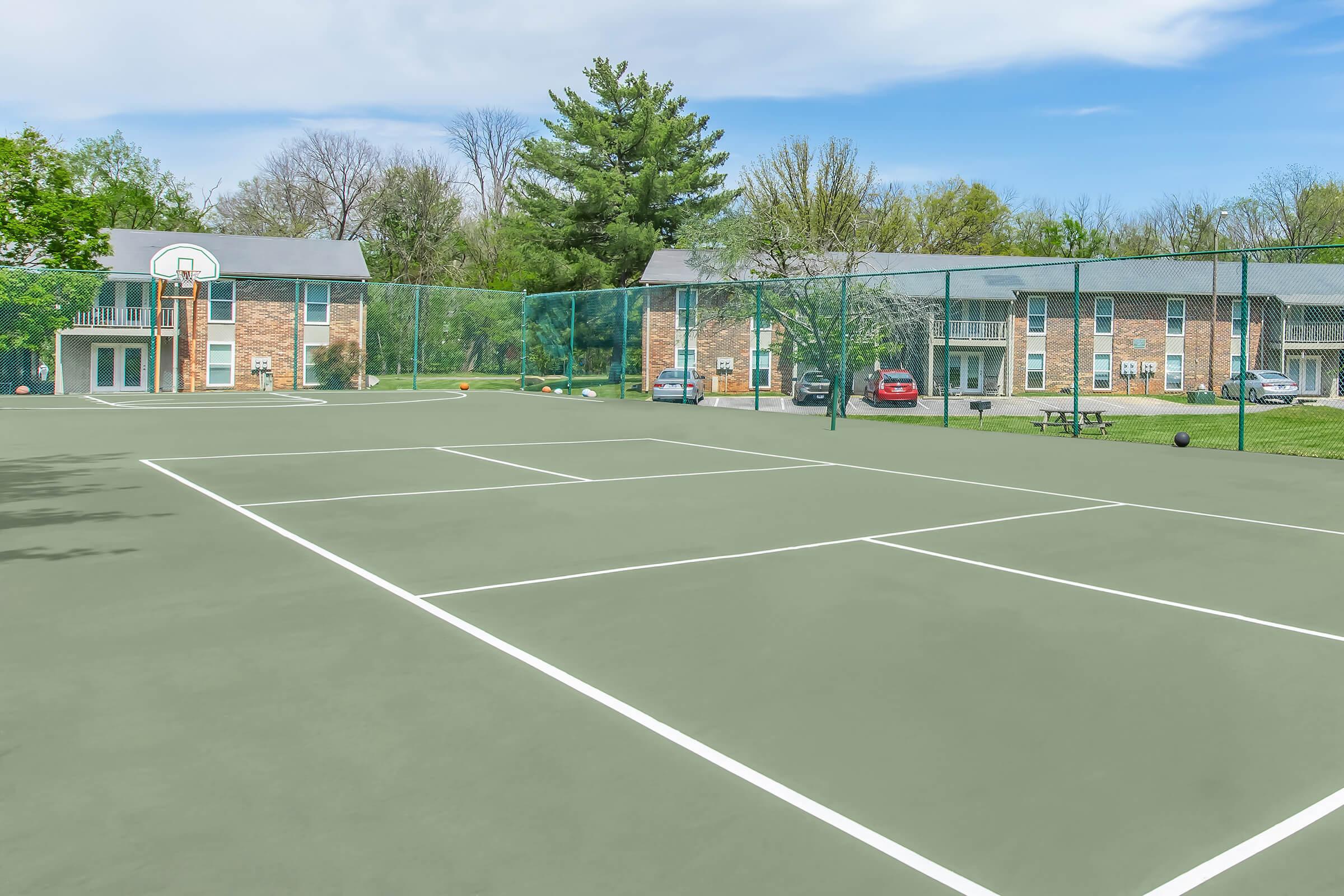 A view of a tennis court with white lines and a basketball hoop in the background. Nearby are residential buildings surrounded by greenery, with a couple of parked cars visible. The sky is clear, creating a bright and inviting atmosphere for recreational activities.