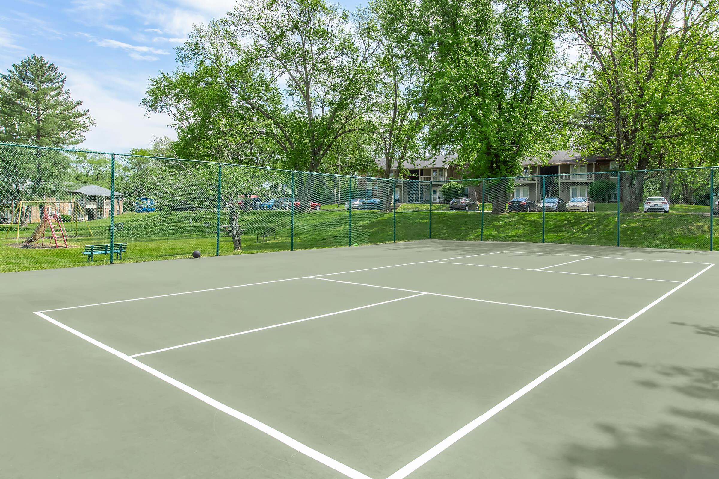 A well-maintained outdoor tennis court surrounded by lush green trees. In the background, there are residential buildings and parked cars, as well as a playground visible to the left. The sky is clear with a few clouds, creating a bright and inviting atmosphere.