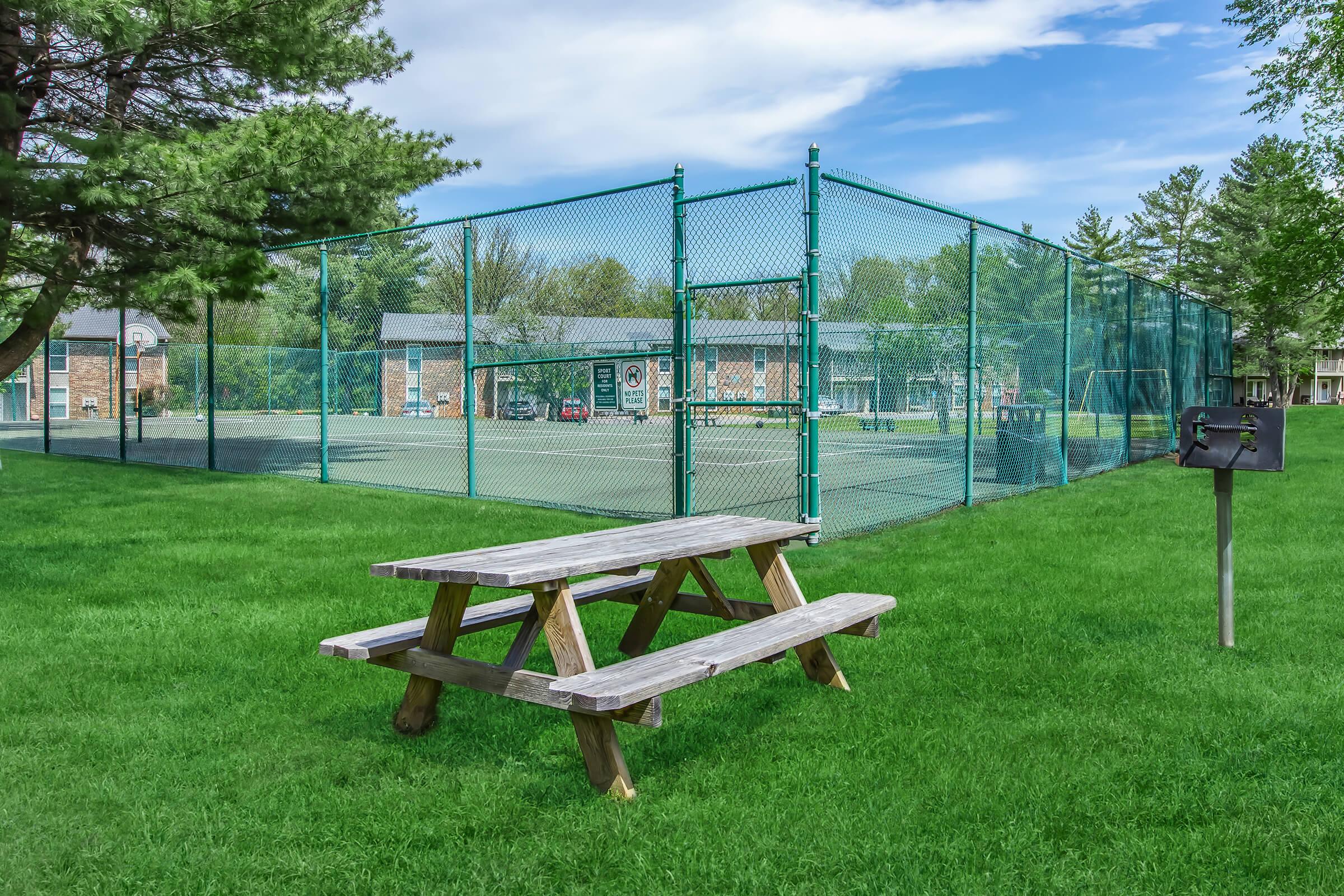 A grassy area featuring a wooden picnic table in the foreground. Behind it, a fenced tennis court and a multi-story building can be seen. The sky is clear with some clouds, and trees provide shade around the area.