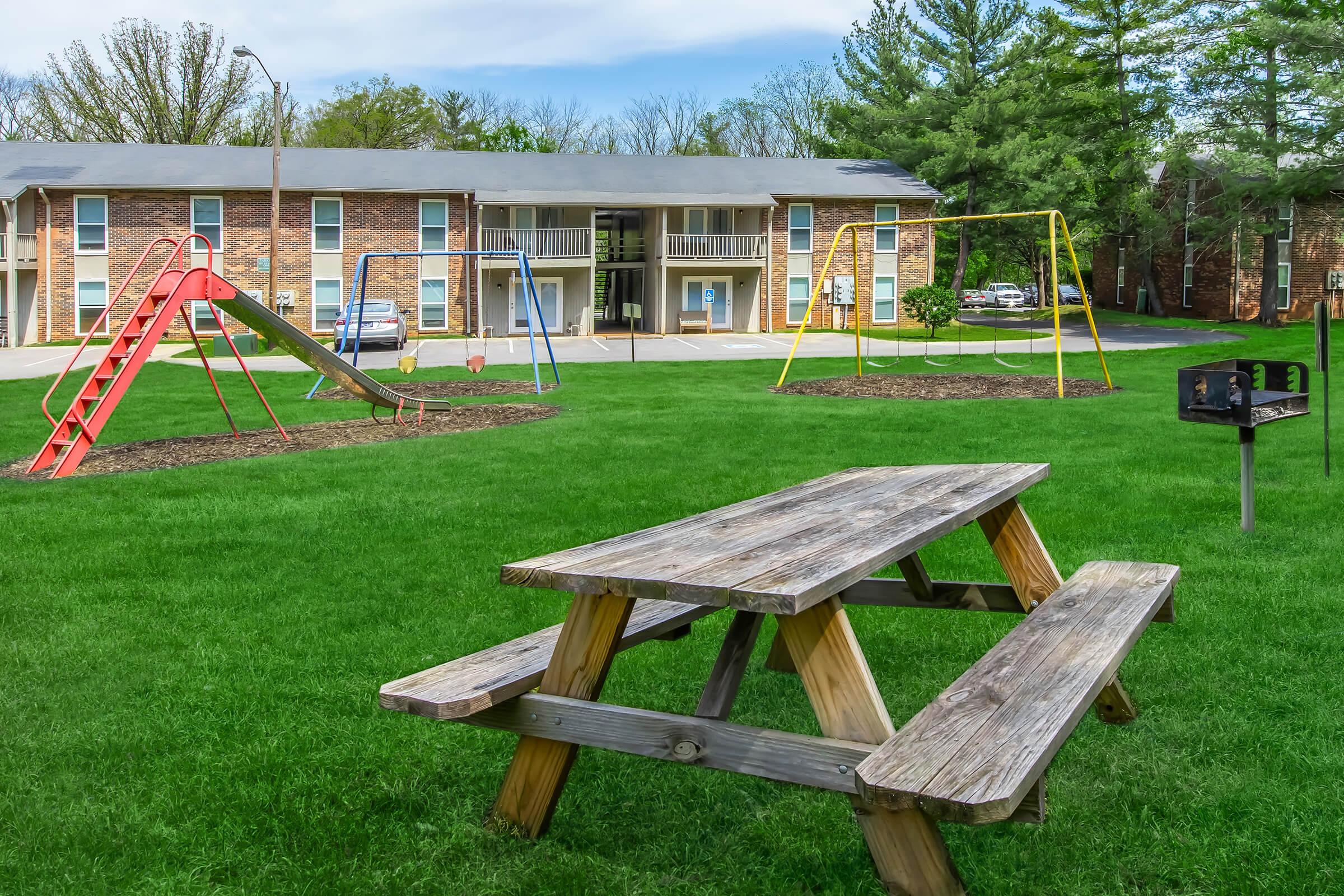 A grassy area featuring a red slide and yellow swings set in front of multiple brick apartment buildings. A wooden picnic table is positioned in the foreground, and there is a charcoal grill nearby. The scene is bright and inviting, with trees in the background.