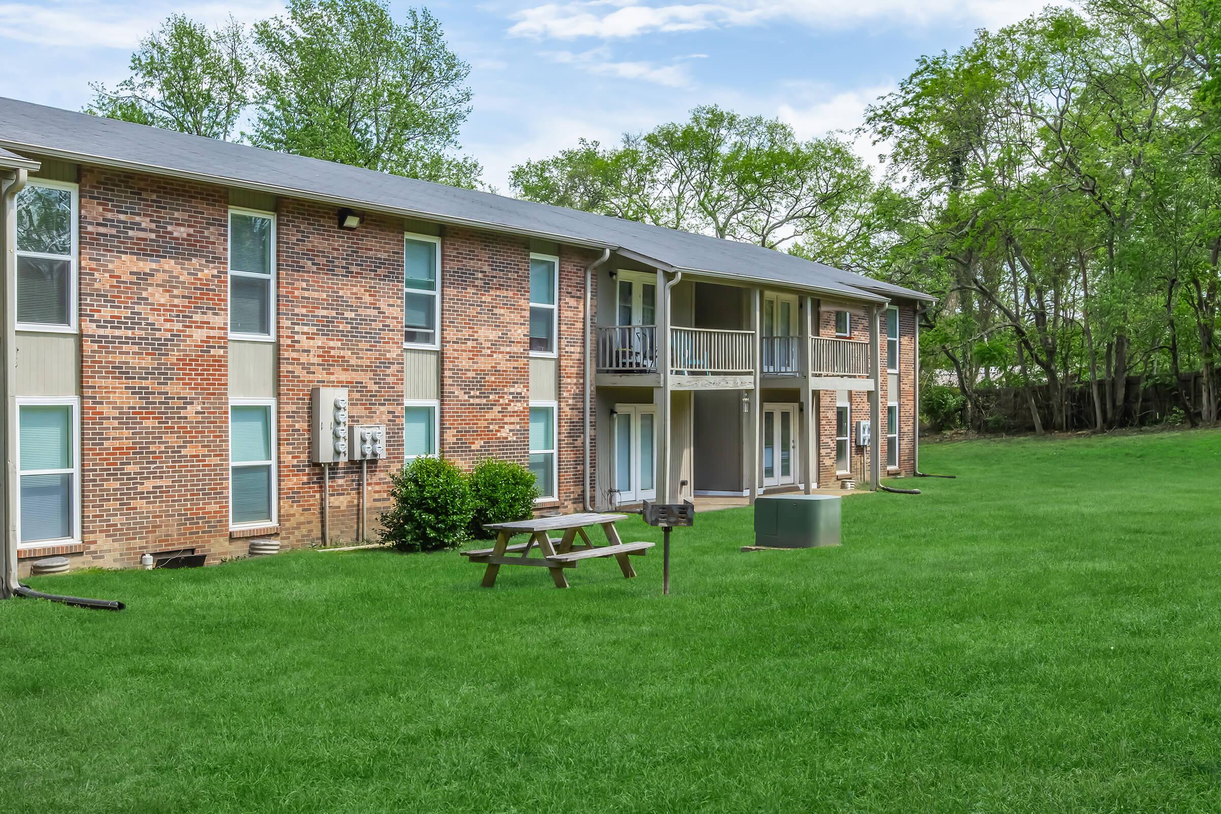 A well-maintained apartment complex featuring brick exterior, multiple units with balconies, and a spacious green lawn. In the foreground, there is a picnic table and a utility box, surrounded by trees and blue skies. Пeaceful residential environment.