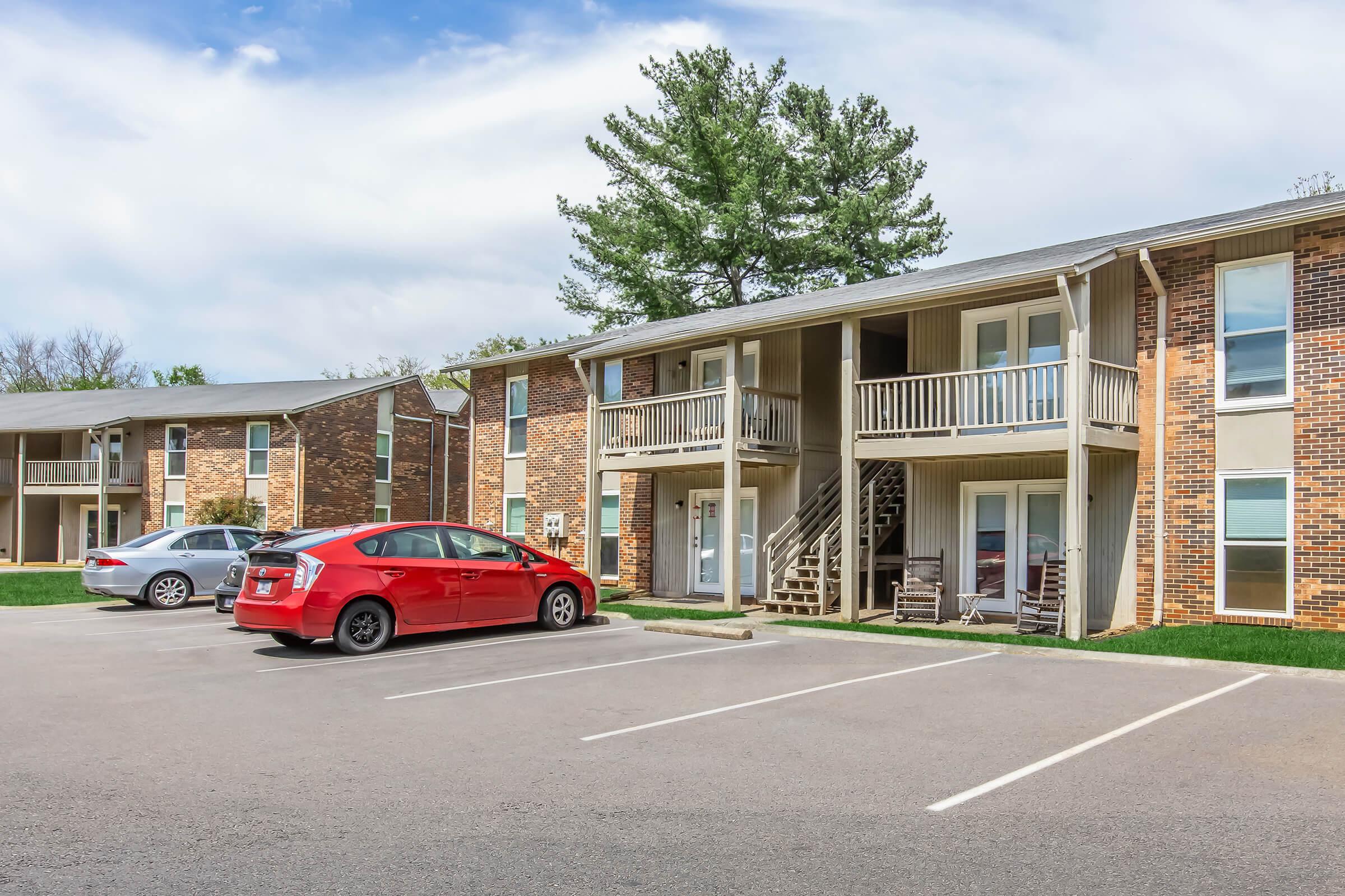 Two-story apartment buildings with brick exteriors. A red car is parked in the foreground, alongside a gray car. Two wooden rocking chairs are visible on a balcony. Green trees in the background and blue sky with clouds above. The area appears well-maintained with a parking lot.