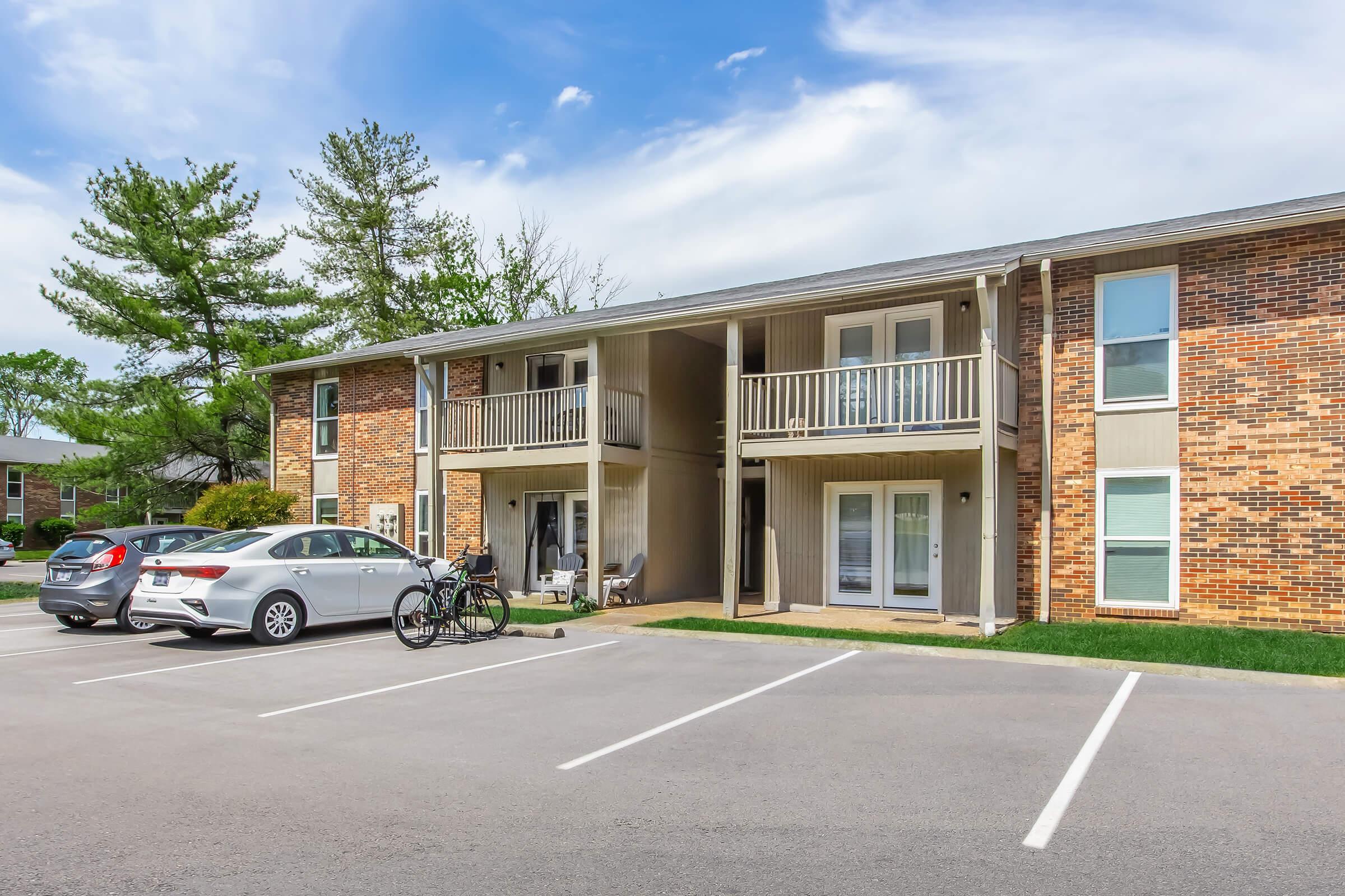 A residential apartment building with two levels, featuring brick and beige siding. Balconies are visible on the upper floor. In the foreground, there are several parked cars and a bicycle. The setting includes grassy areas and trees under a partly cloudy sky.