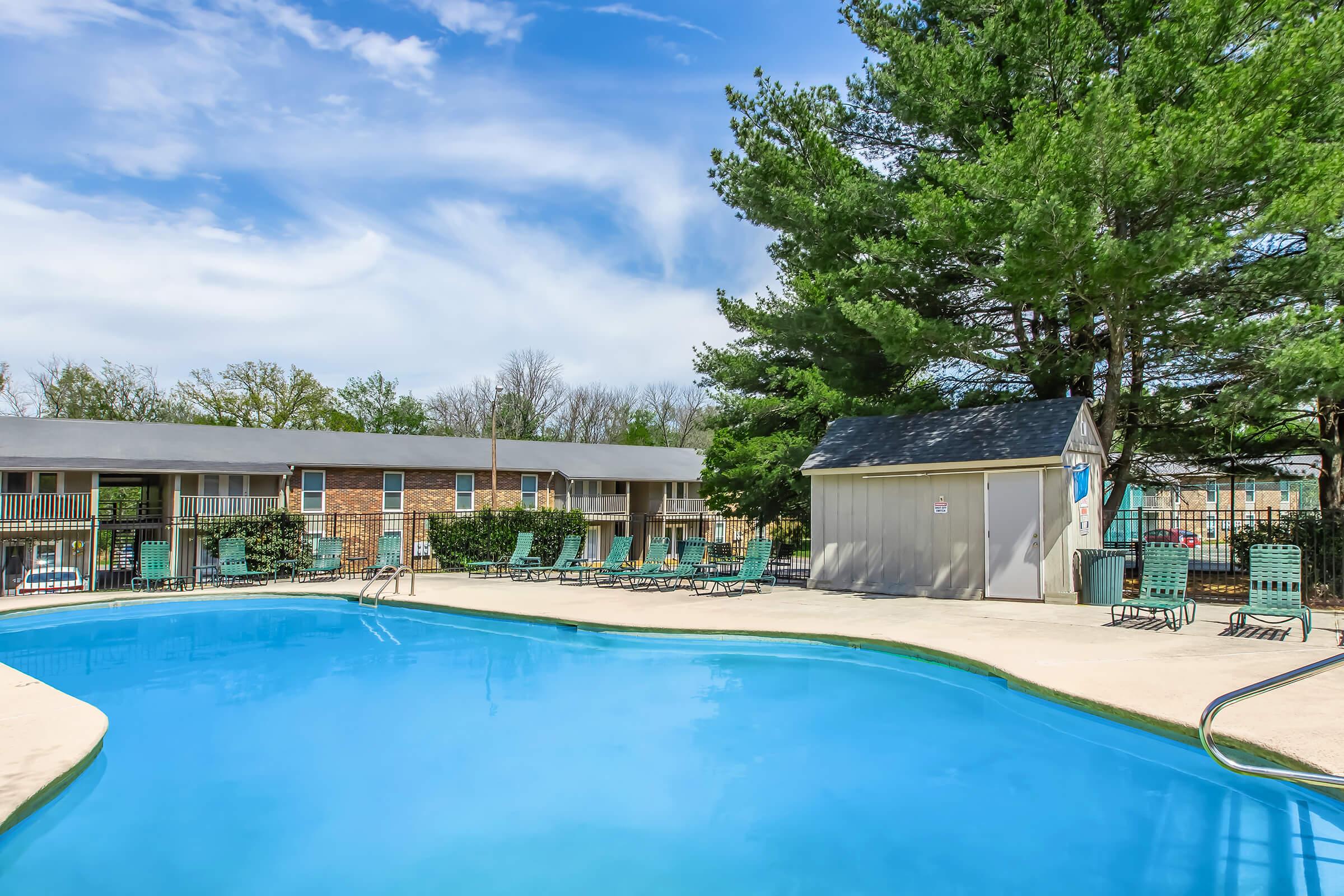 A clear blue swimming pool surrounded by lounge chairs, with a small shed nearby. Lush green trees are visible in the background, and a bright blue sky features scattered clouds. The setting suggests a relaxing outdoor space, likely at an apartment complex or recreational area.