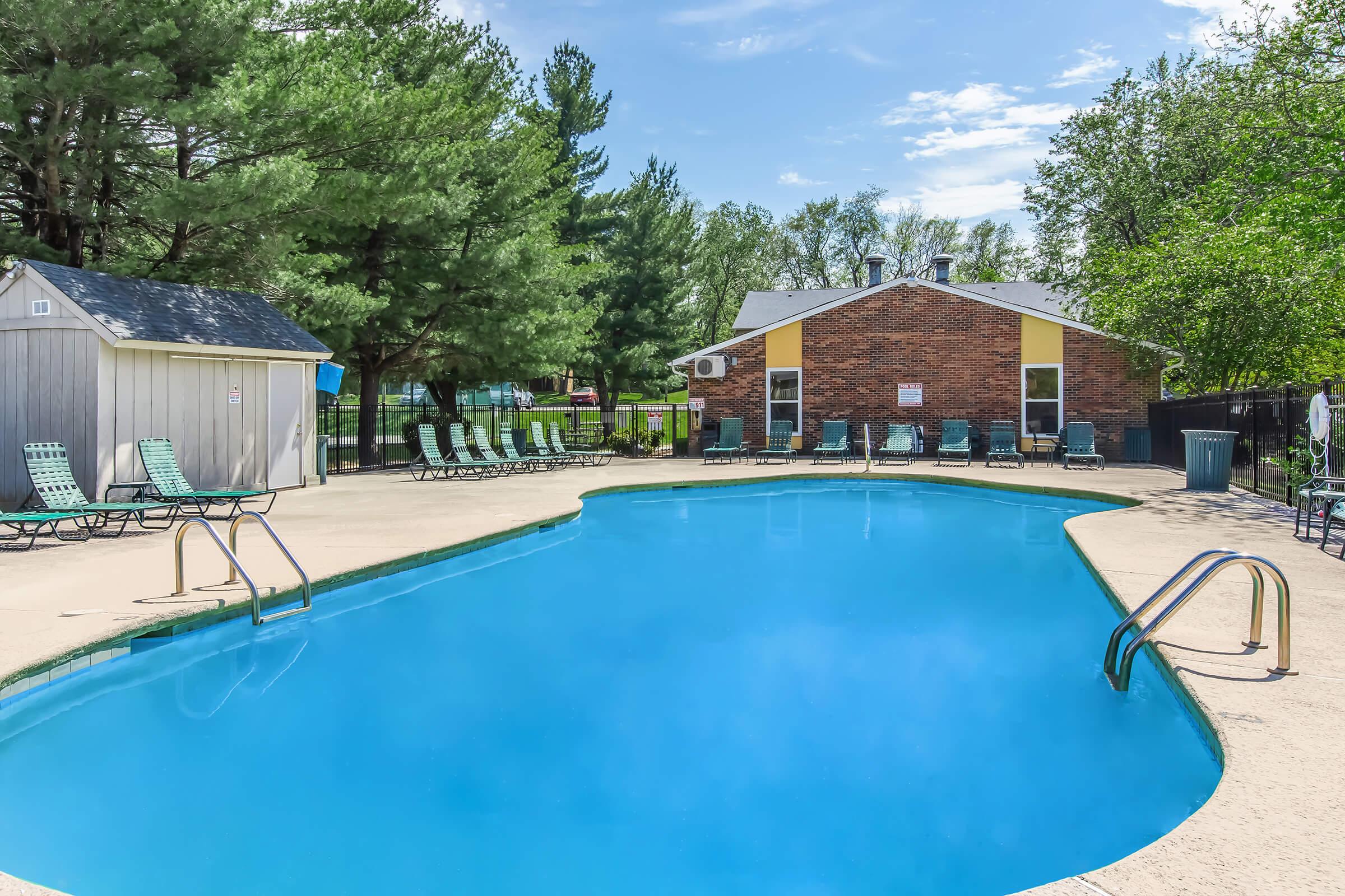 A clear blue swimming pool surrounded by lounge chairs, with trees in the background and a brick building nearby. The scene is set on a sunny day, showcasing a peaceful outdoor recreational area.