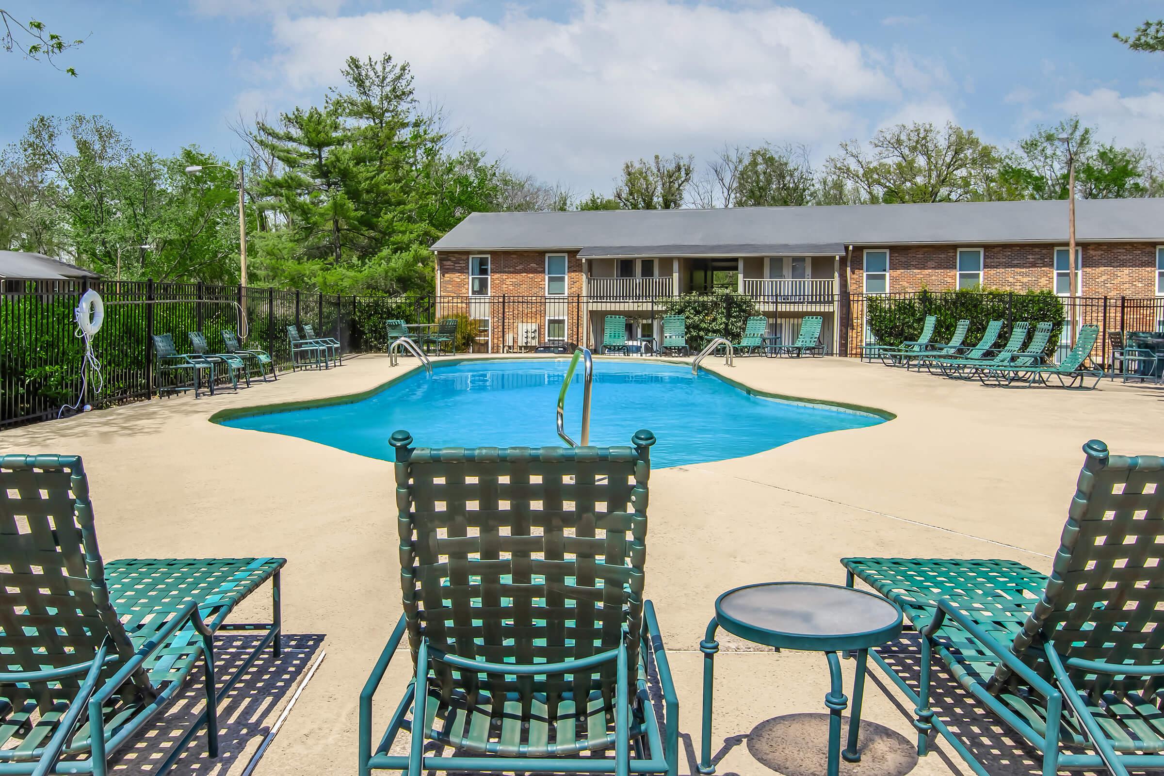 A clear swimming pool surrounded by a patio area with green lounge chairs. Trees and a building with balconies are visible in the background under a partly cloudy sky. The setting appears serene and inviting, ideal for relaxation.