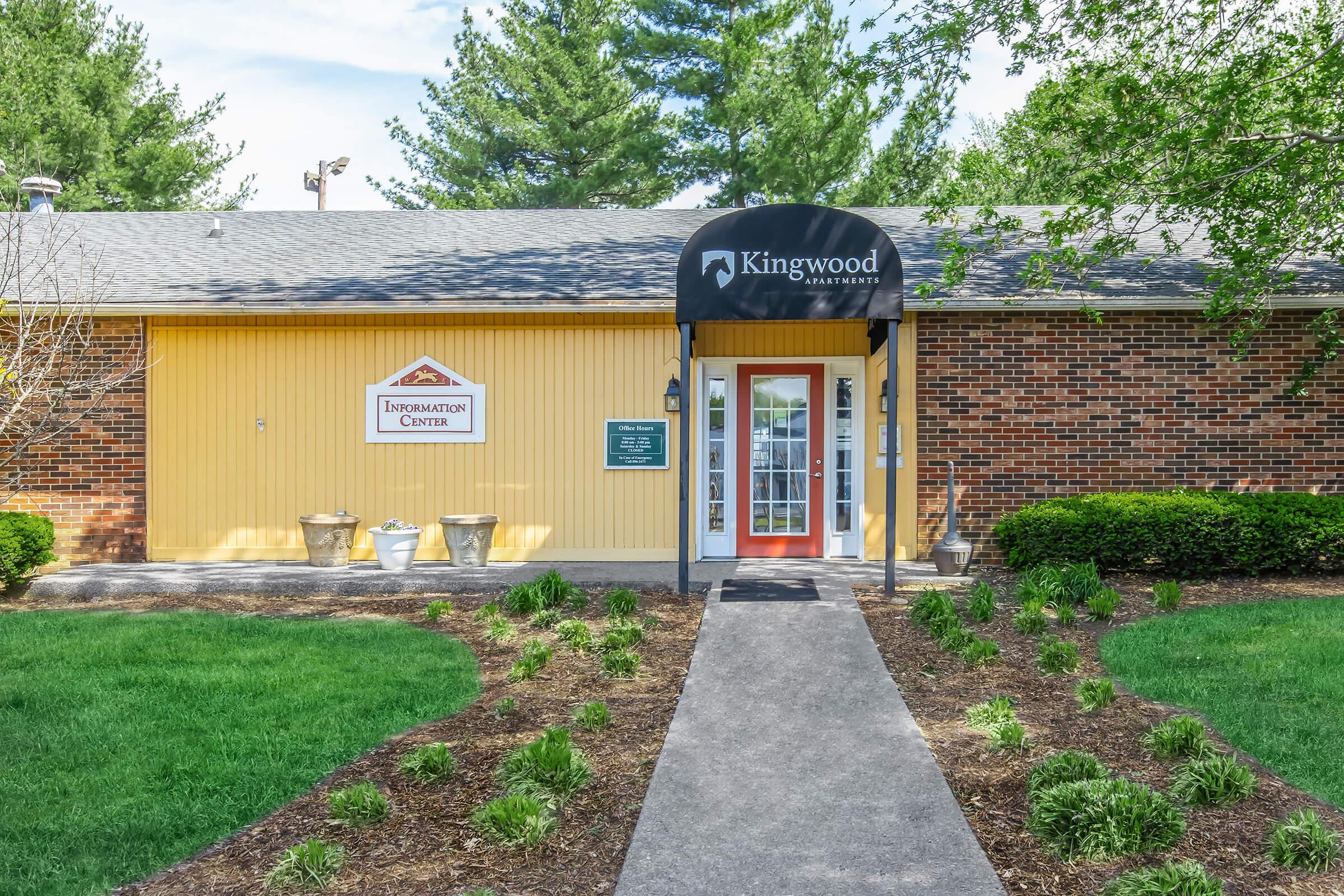 Entrance to the Kingwood Community Center, featuring a covered porch with glass door and large windows. The exterior is brick with yellow accents, bordered by green lawns and landscaped flower beds. A sign indicates the building is the Information Center. Trees and shrubs add to the pleasant surroundings.