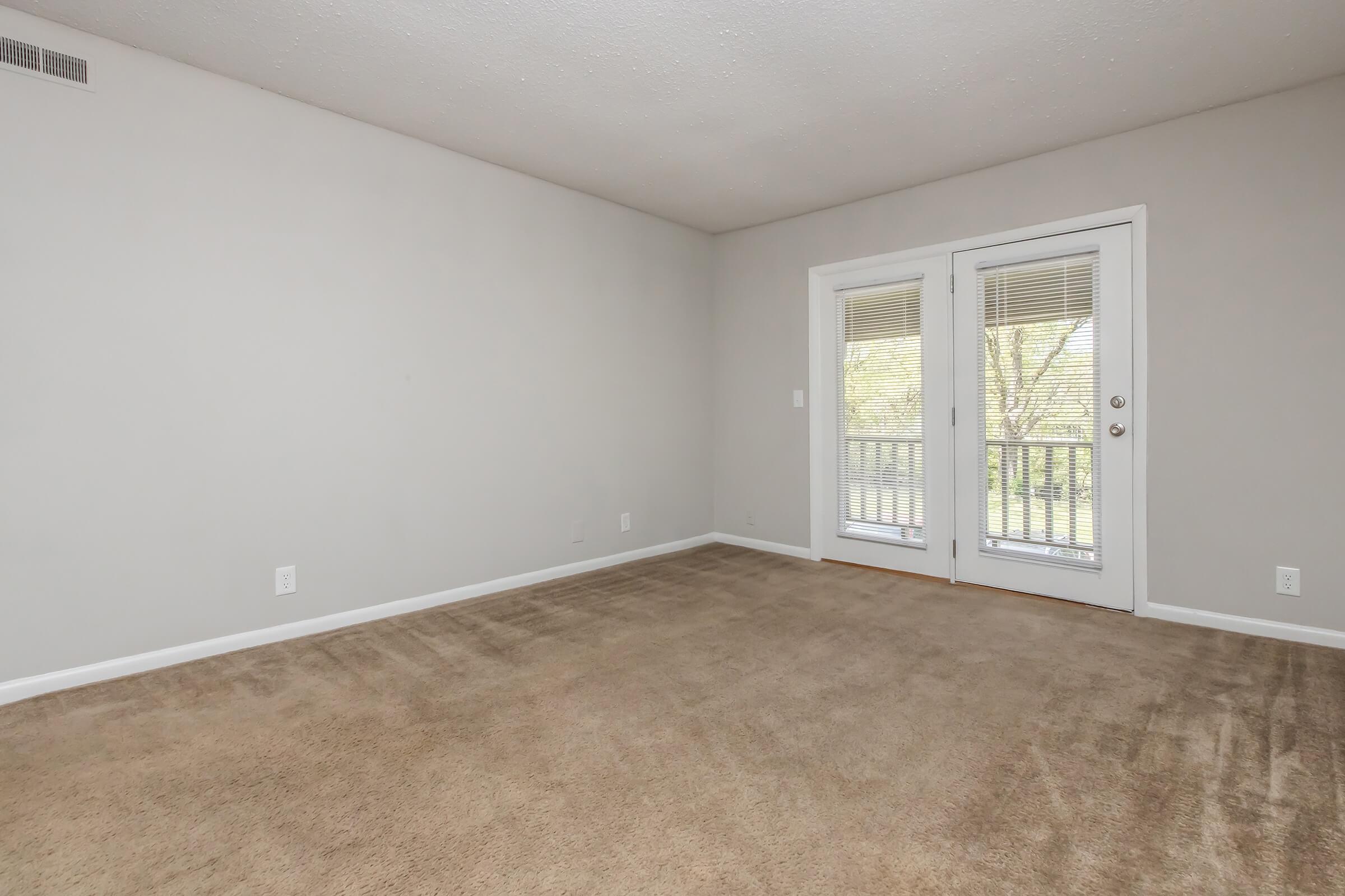 A vacant room featuring light gray walls and beige carpet. It has a set of double doors leading to a balcony with natural light coming in. The space is minimalistic with no furniture, emphasizing the open floor plan and clean, empty atmosphere.