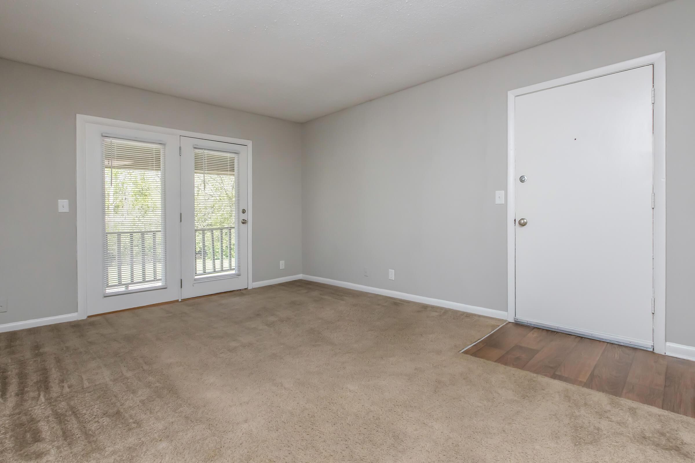 A spacious, empty living room featuring light gray walls and a plush beige carpet. The room has a set of double glass doors leading to a balcony, along with a single front door. Natural light filters in, creating a bright and welcoming atmosphere.
