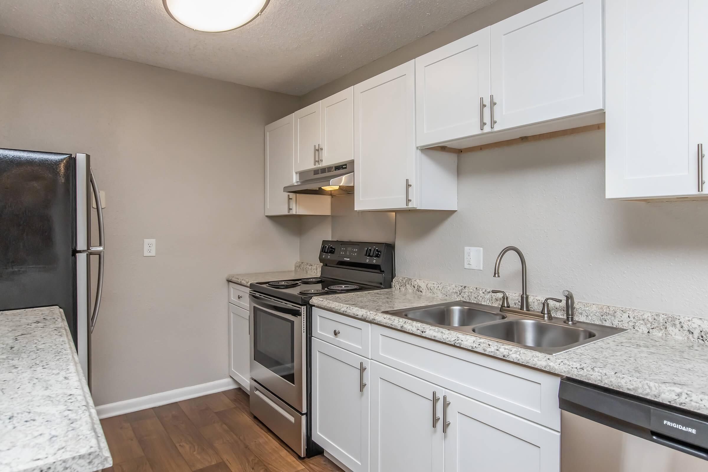 Modern kitchen featuring white cabinetry, stainless steel appliances, a granite countertop, and a double sink. The space includes an oven, stove, and microwave, with a neutral wall color and hardwood flooring. Bright overhead lighting enhances the contemporary design.