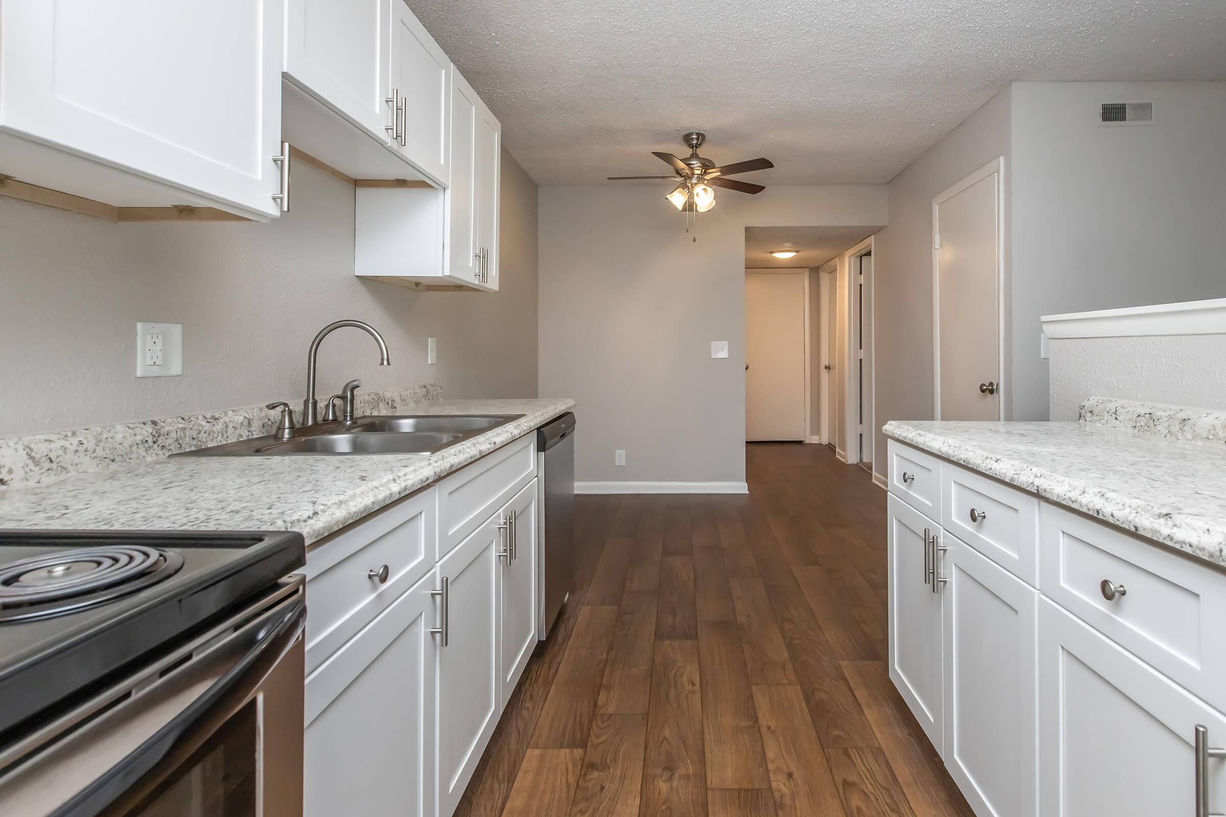 Modern kitchen with white cabinets, granite countertops, stainless steel appliances, and a ceiling fan. The kitchen area is open and well-lit, leading to a hallway that connects to other rooms. The wood-like flooring adds warmth to the space.