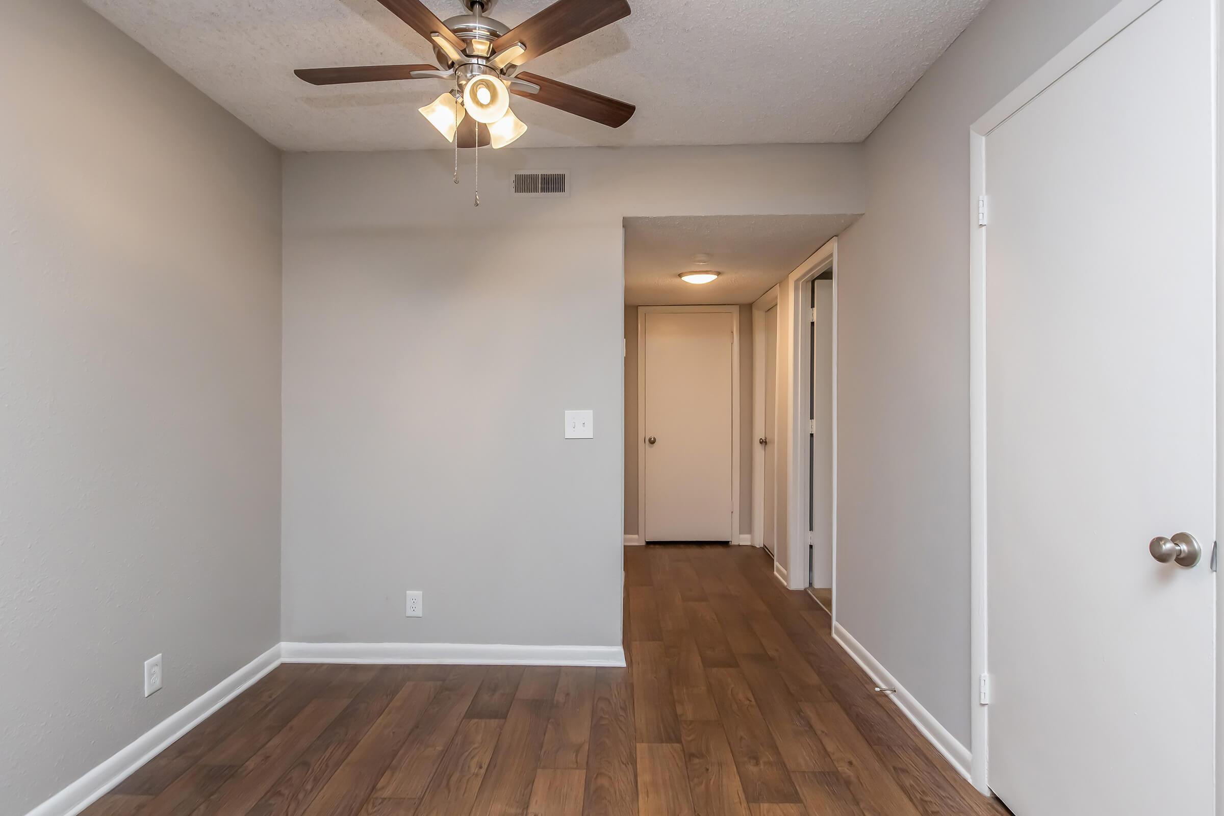 A well-lit room featuring a ceiling fan, light-colored walls, and laminate flooring. The room includes a door leading to a hallway on the right and another closed door in the background. The space has a clean, modern aesthetic suitable for various uses.