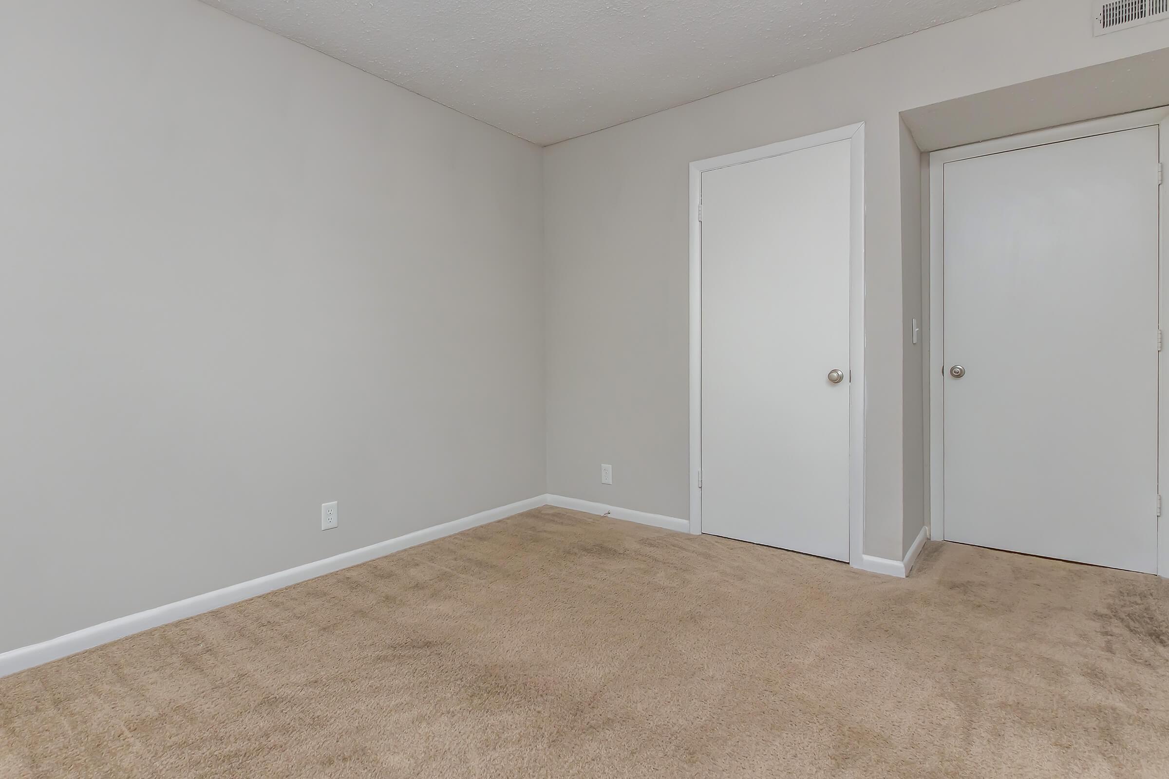 An empty room with beige carpet and light gray walls. One door is closed, leading to an unknown area, while another door on the right side is also closed. The space is bright and uncluttered, creating a minimalist atmosphere.