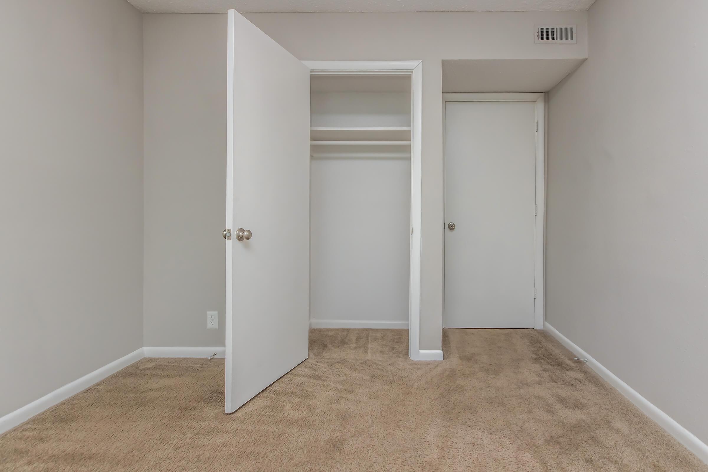 An empty room with beige carpet and light gray walls. There are two doors: one leading to a closet with a single shelf and the other to another small space. The room has no furniture or decorations, giving it a clean and minimal look. Natural light filters in from an unseen source.