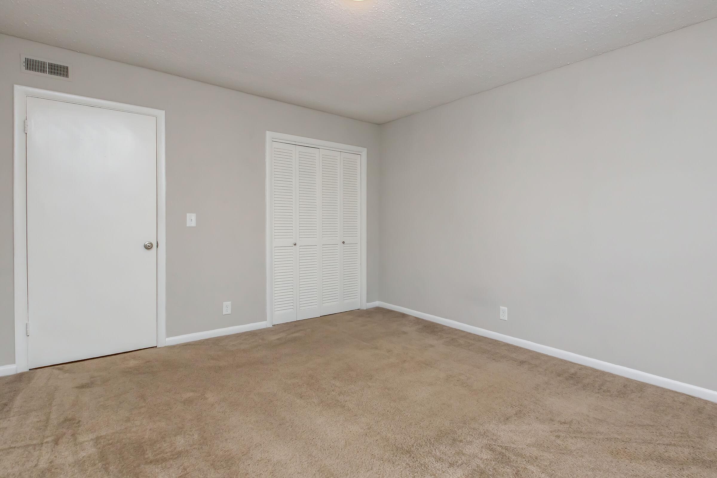A plain empty room with beige carpet, light gray walls, and a white door on the left. There is a closet with white bi-fold doors opposite the door, and no furniture or decorations are visible. The space is well-lit with natural light coming in.