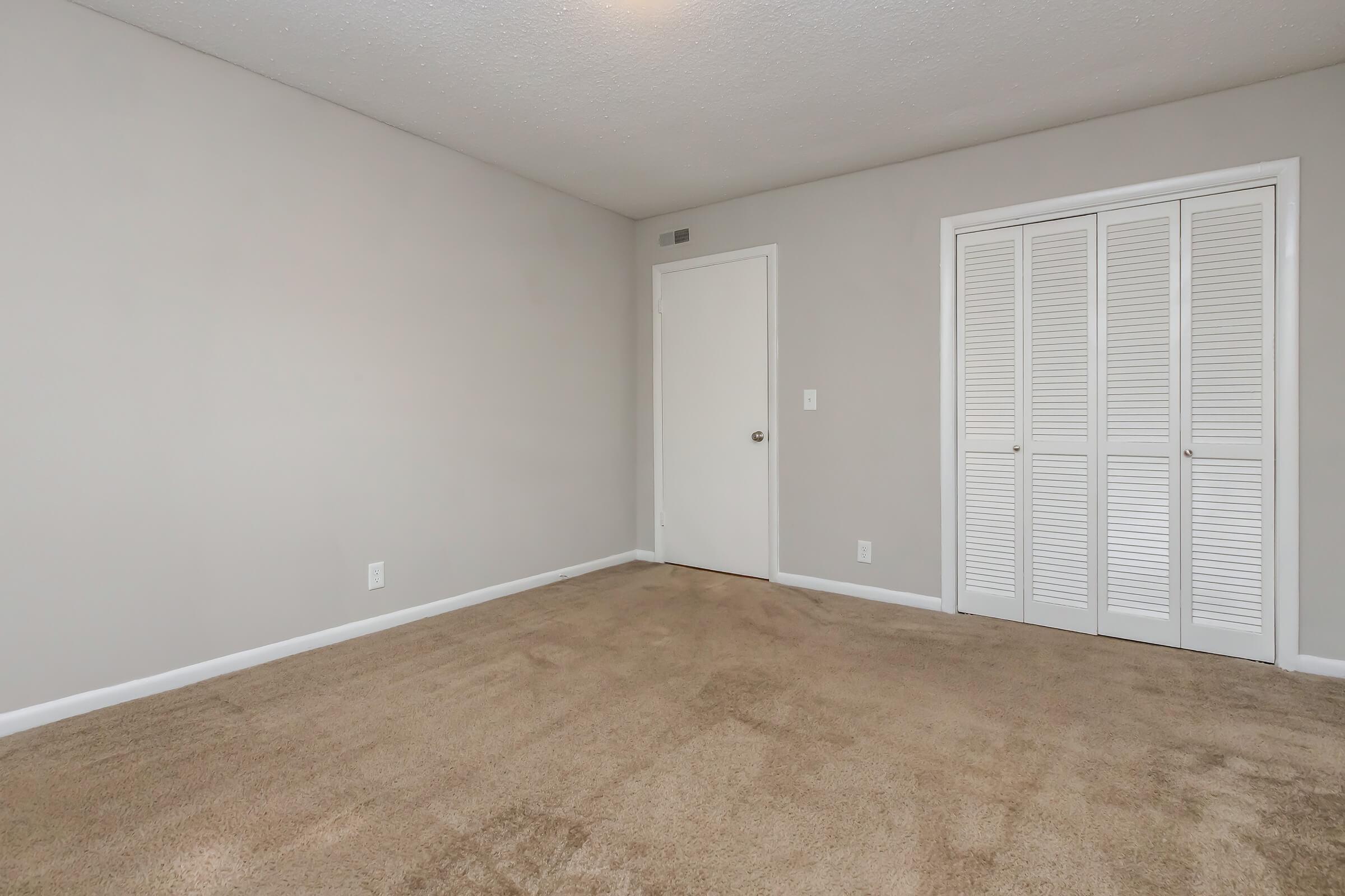 Empty room with beige carpet and light gray walls, featuring a closed white door and a closet with louvered doors. The space is well-lit, giving a spacious feel, and has no furniture or decorations.