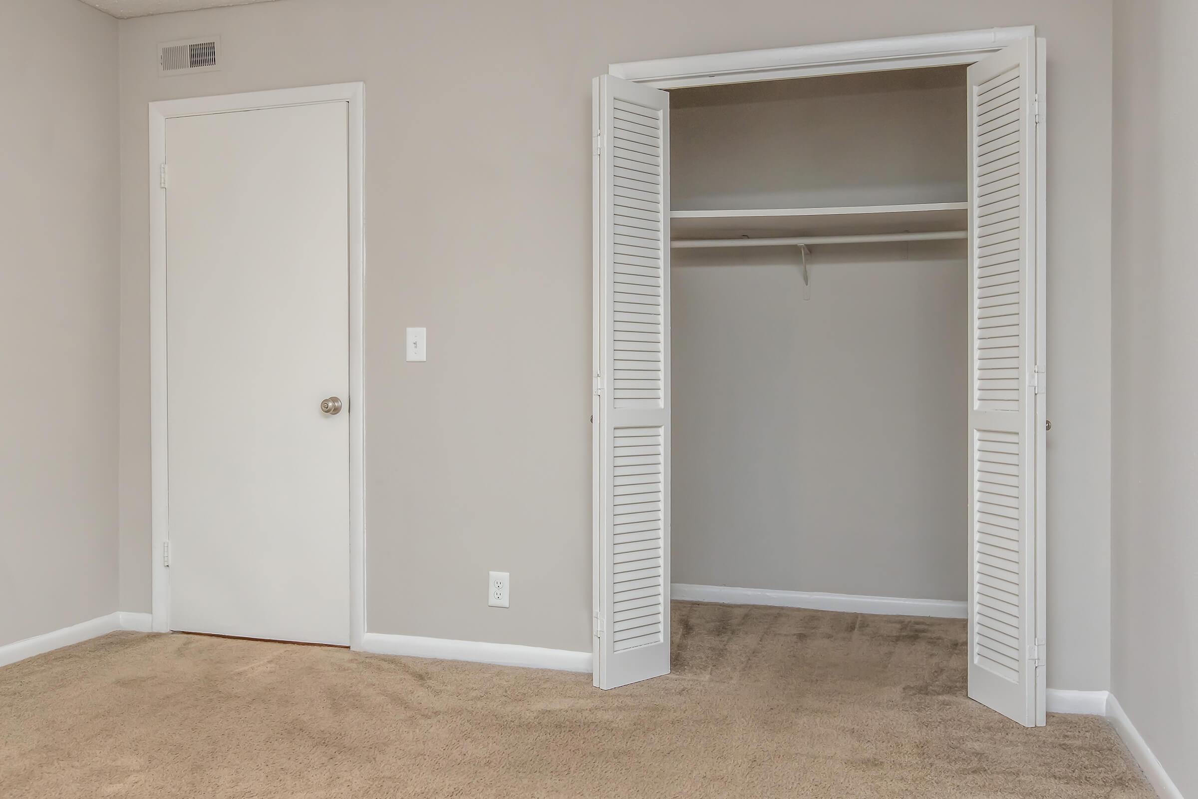 A partially empty room with beige carpet and light gray walls. A closet with white louvered doors is to the right, showing a hanging rod inside. A closed white door is visible on the left side of the image. The overall atmosphere is clean and minimalistic.