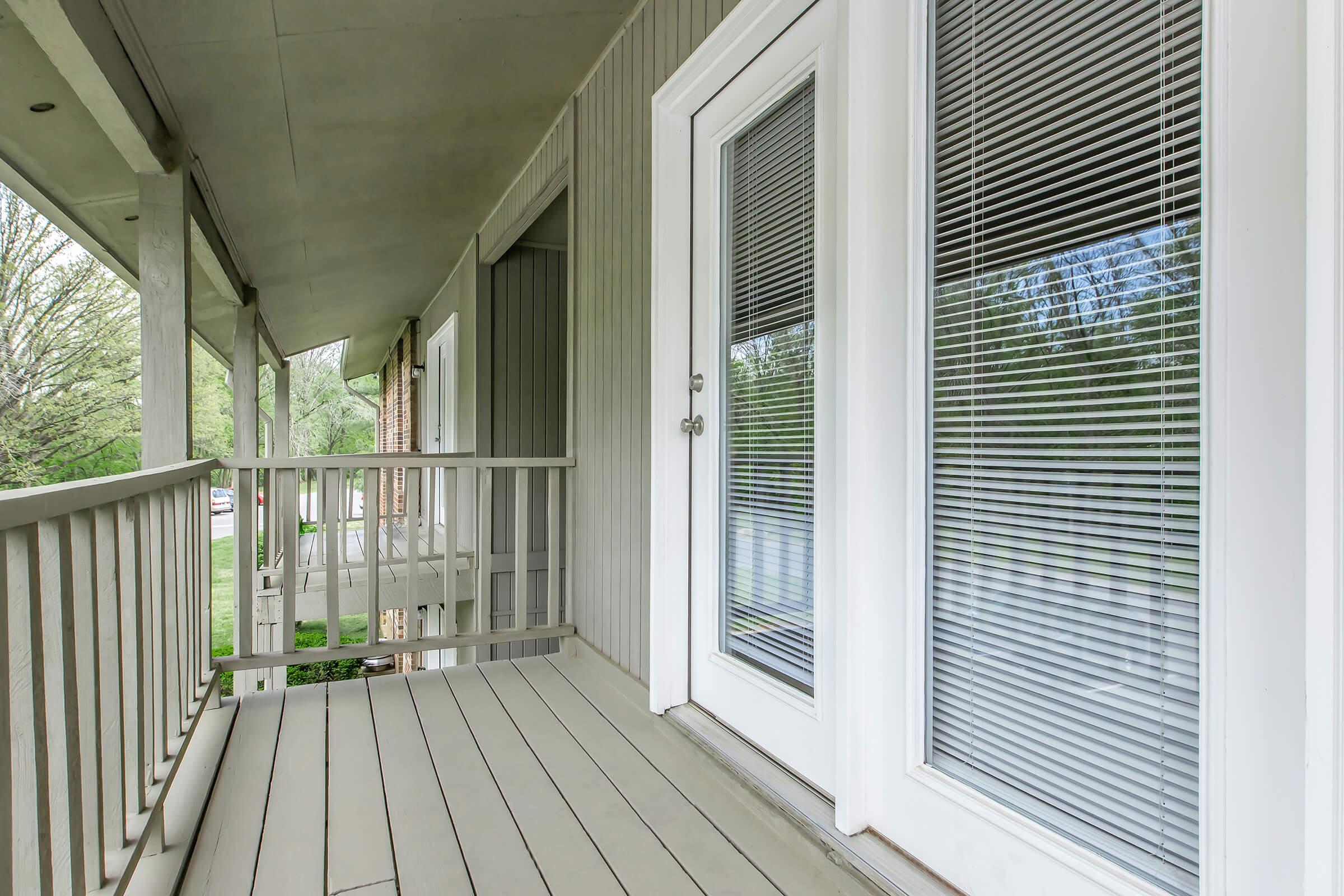 A close-up view of a porch with wooden flooring and a white door featuring blinds. The porch has a railing and overlooks a green landscape. Soft natural light filters through the blinds, highlighting the clean lines and modern design of the space.
