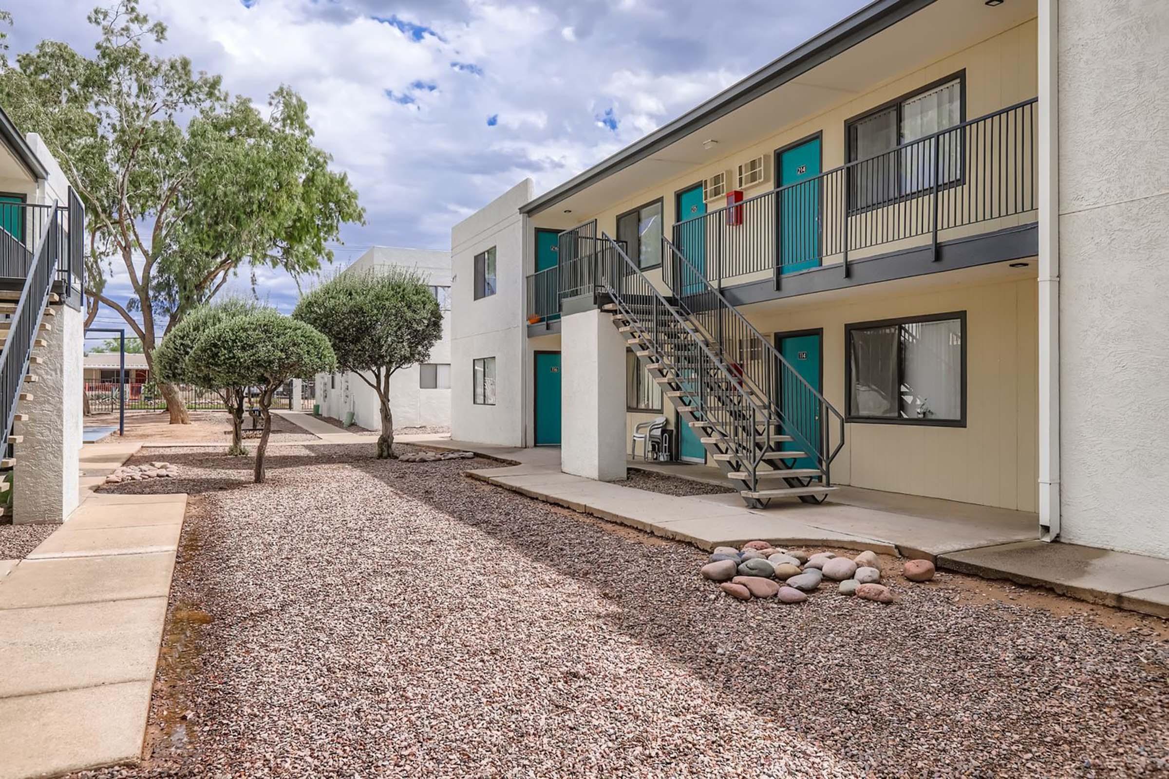 View of a multi-unit apartment building with two stories. The exterior features a minimalist design with light-colored walls and teal doors. A staircase leads to the second floor, and there are landscaped areas with small trees and gravel paths. The sky is partly cloudy, adding a pleasant atmosphere to the scene.
