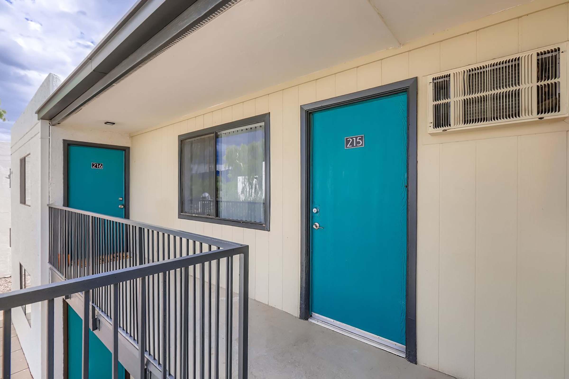 Two hotel room doors, one labeled "214" and the other "215", both painted in a vibrant turquoise color. The doors are set in a light-colored building with a balcony railing, and there is an air conditioning unit mounted above the door to room 215. The sky is partly cloudy, providing a serene atmosphere.