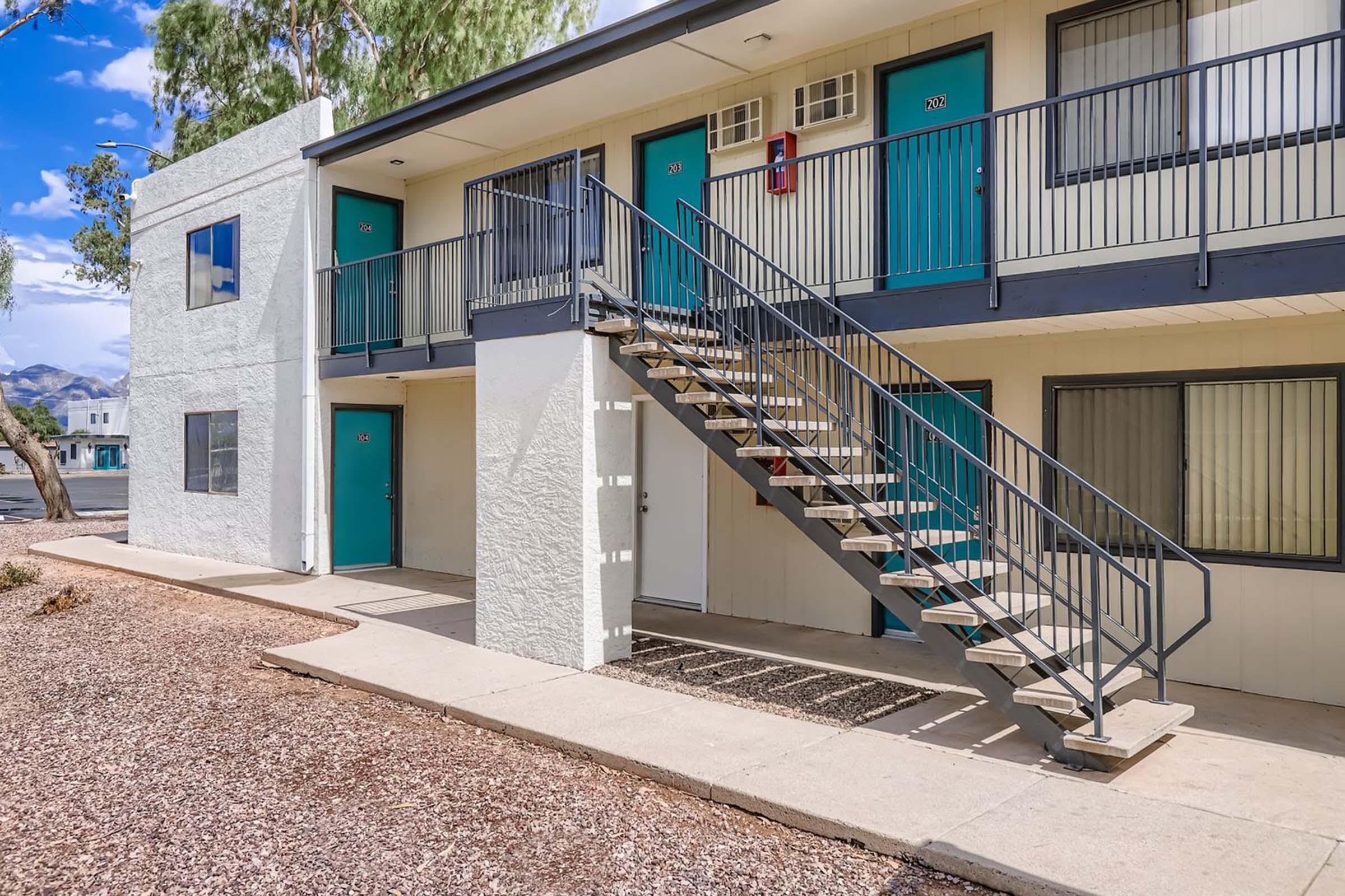 Exterior view of a two-story apartment building with a set of stairs leading to the upper floor. The building has light-colored walls with turquoise doors. The surrounding area features gravel landscaping and a few trees, with a clear blue sky in the background.