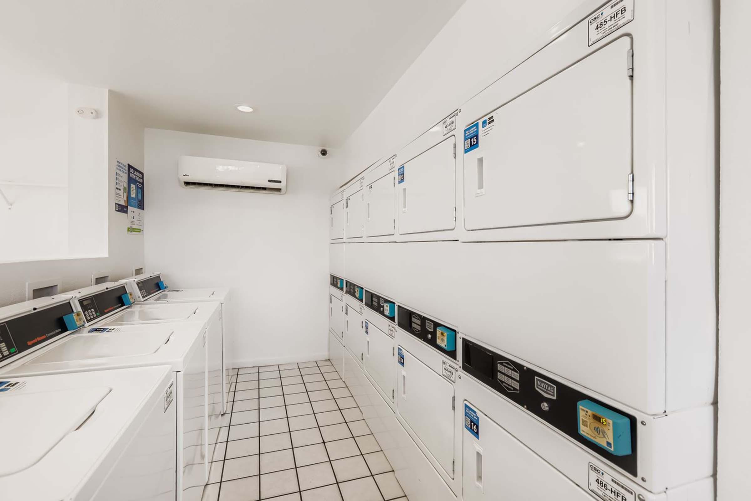 A clean, well-lit laundry room featuring rows of white washing machines and dryers with digital controls. The space has white tiled flooring and a climate control unit on the wall, creating a bright and functional environment for laundry tasks.