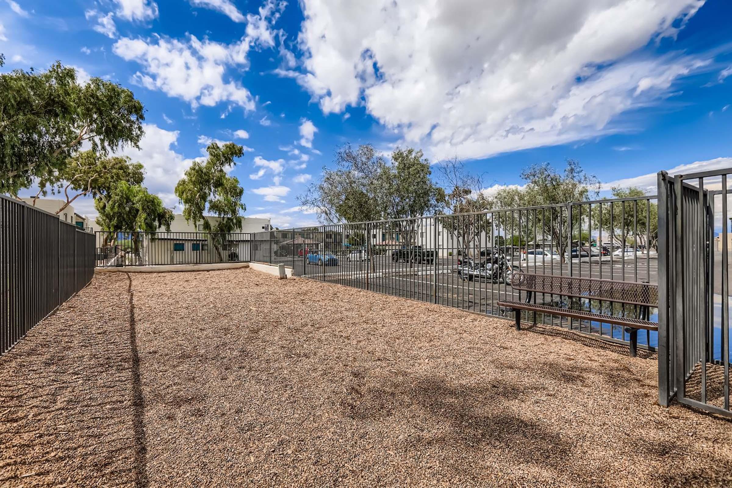 A fenced outdoor area with a gravel surface and a bench, surrounded by trees under a partly cloudy sky. In the background, there are buildings and a parking lot. The setting appears to be a calm, recreational space.